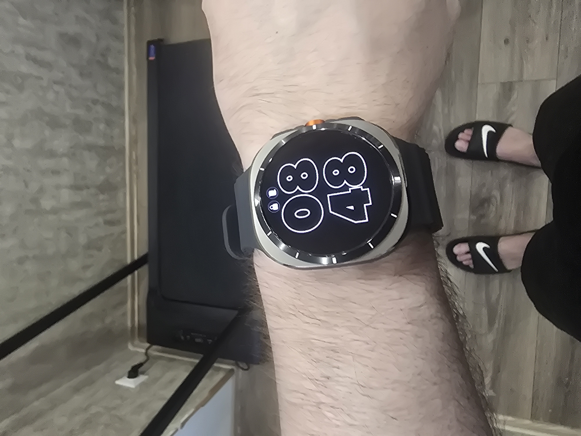 A first-person perspective view of a person's left arm wearing a smartwatch displaying 08:48. The person is also wearing black Nike slides and black pants. In the background, a black treadmill is visible, and a white electrical outlet with a plug is mounted on a grey, textured wall. The flooring appears to be light-colored wood laminate.