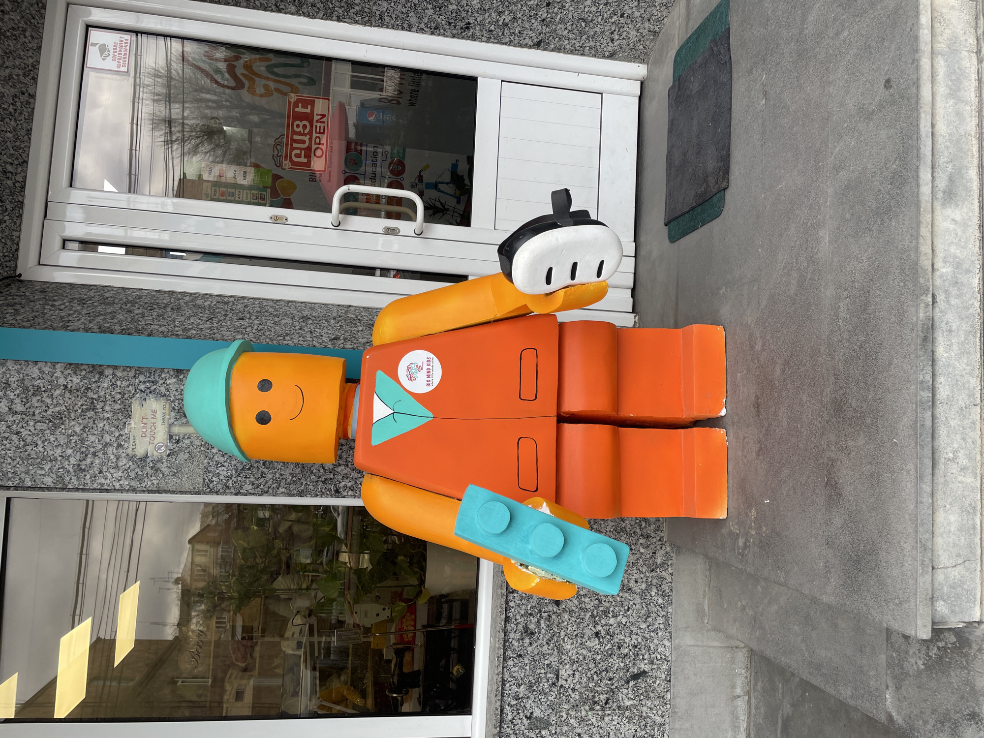 The image shows a life-sized Lego-style figure positioned in front of a building's entrance. The figure, primarily orange, has a teal hard hat and is holding a white object resembling a headset in one hand and a blue Lego brick in the other. It has a simple, painted-on smile. A sticker with the logo of "Big Mind Kids" is visible on its chest. The background includes the entrance of a building with a "PUSH OPEN" sign in both Armenian and English, along with visa and other education advertisements. A small sign reading "PLEASE DON'T TOUCH ME THANK YOU" is visible near the figure's head. The location is the city of Yerevan, Armenia. The building appears to be a business or educational facility.
