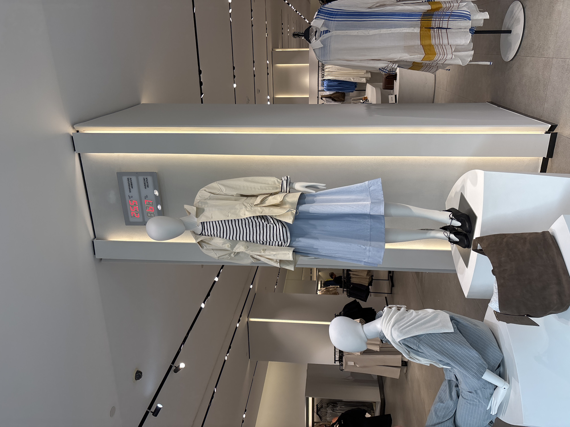 The image shows a retail clothing store interior. In the foreground, two mannequins are displayed. The mannequin on the right is standing and wears a striped t-shirt, a light-colored jacket, and a light blue skirt. It is wearing black shoes. A digital display showing temperature and humidity is mounted on the wall behind it. The mannequin on the left is posed sitting down, wearing a striped grey outfit and a white sweater draped around its shoulders. A brown suede handbag is placed on a white display pedestal in front of it. In the background, clothing racks with various garments are visible, along with more display areas. The overall environment appears to be a well-lit, modern retail space. There is no visible indication that this scene is located in the city of Orihuela, Spain.