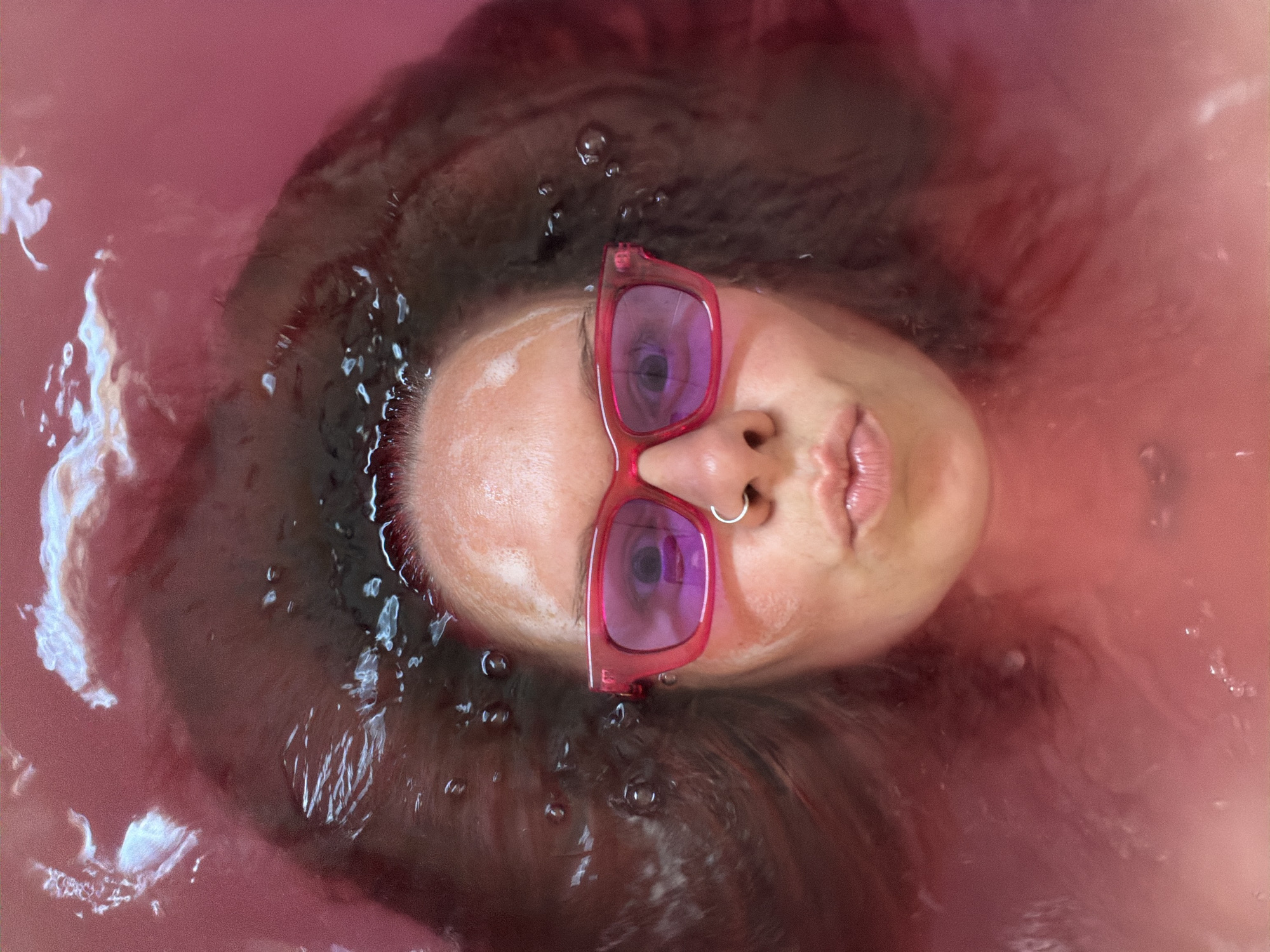 This is an overhead, close-up, eye-level shot of a person in a pink liquid, possibly a bath. The person is wearing bright pink sunglasses and has a silver hoop nose ring. Their hair is dark brown and appears wet, fanned out around their head. The pink liquid is murky and has some bubbles on the surface, with highlights suggesting light reflecting off of it. The person's lips are puckered, and they are looking towards the camera. The image does not contain any specific information about the city of Chaikovsky, Russia.