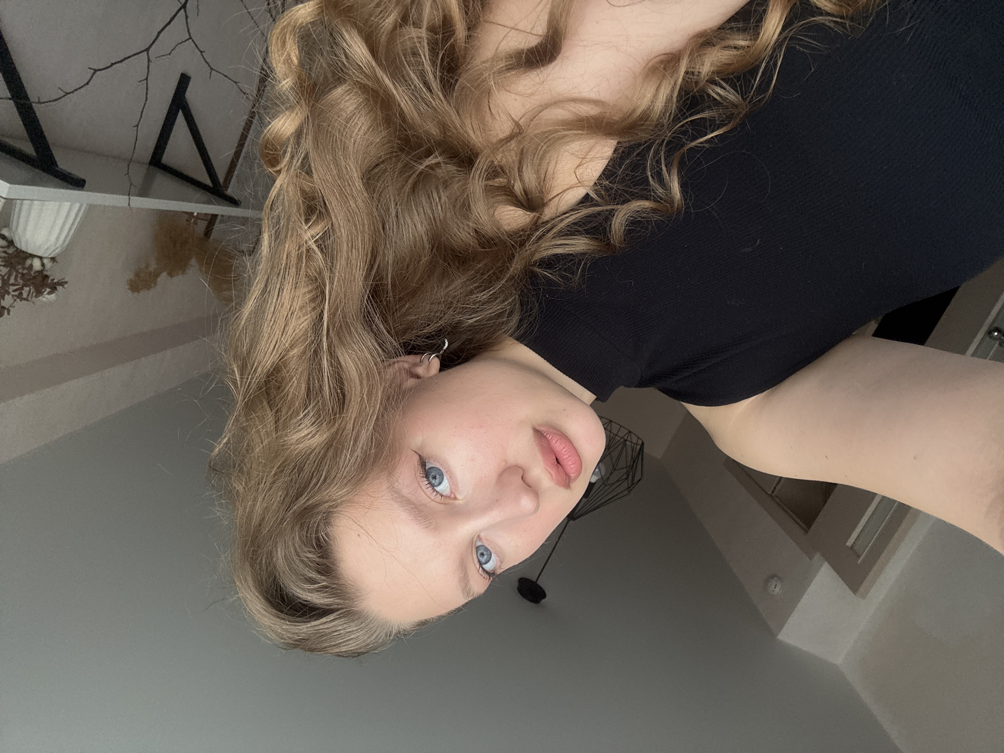 The image is a selfie taken indoors, likely in a residential setting. A young woman with blonde, wavy hair and blue eyes is the primary subject. She is wearing a black, high-necked top. Her makeup includes winged eyeliner and lipstick. She has a small hoop earring in her visible ear. The background shows a ceiling with a light fixture and what appears to be a doorway. A shelf with decorative items, including dried plants and a vase, is visible in the upper right corner. The overall lighting suggests it is daytime. There is no specific information that would allow for the identification of the city as Izhevsk, Russia.