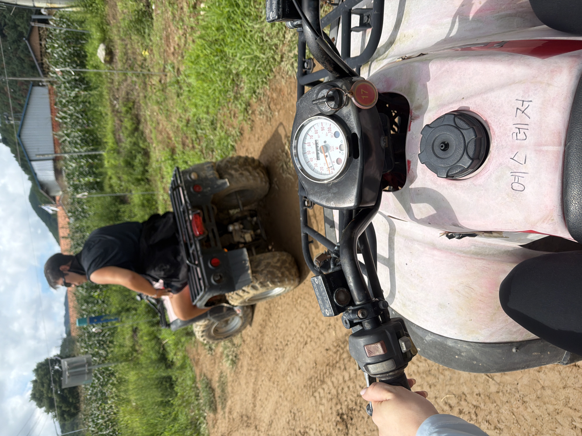In Seo-myeon, South Korea, on a dirt path lined with green foliage and distant buildings, two individuals are riding all-terrain vehicles (ATVs). The person in the foreground is operating a pink ATV, their left hand gripping the handlebars. The ATV features a dashboard with a speedometer, an ignition key with a red tag labeled "17," and a black fuel cap. Handwriting in black marker is visible on the ATV's front panel, reading "예스 레저" (Yes Leisure). The second individual, wearing sunglasses and a black vest over a black shirt and shorts, is riding a black ATV ahead of the first. The environment suggests a rural or recreational setting, possibly a trail for ATV riding, with overcast skies.