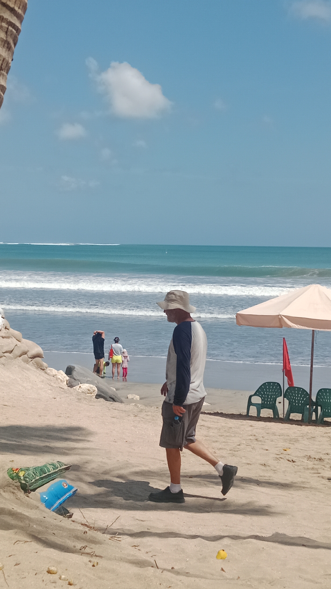 In Kuta, Indonesia, on a sunny day, a man wearing a wide-brimmed hat, a long-sleeved shirt with dark sleeves and a white torso, and grey shorts walks on a sandy beach. He is holding a dark-colored water bottle in his right hand. The beach is populated with several other individuals, including a group of three people, two adults and a child, standing closer to the ocean. In the background, the ocean displays several rows of white waves against a turquoise sea under a bright blue sky with a few scattered clouds. A light-colored umbrella shades two green plastic chairs on the beach. In the foreground, closer to the viewer, a deflated green inflatable toy lies on the sand next to a deflated blue inflatable toy shaped like a dolphin.