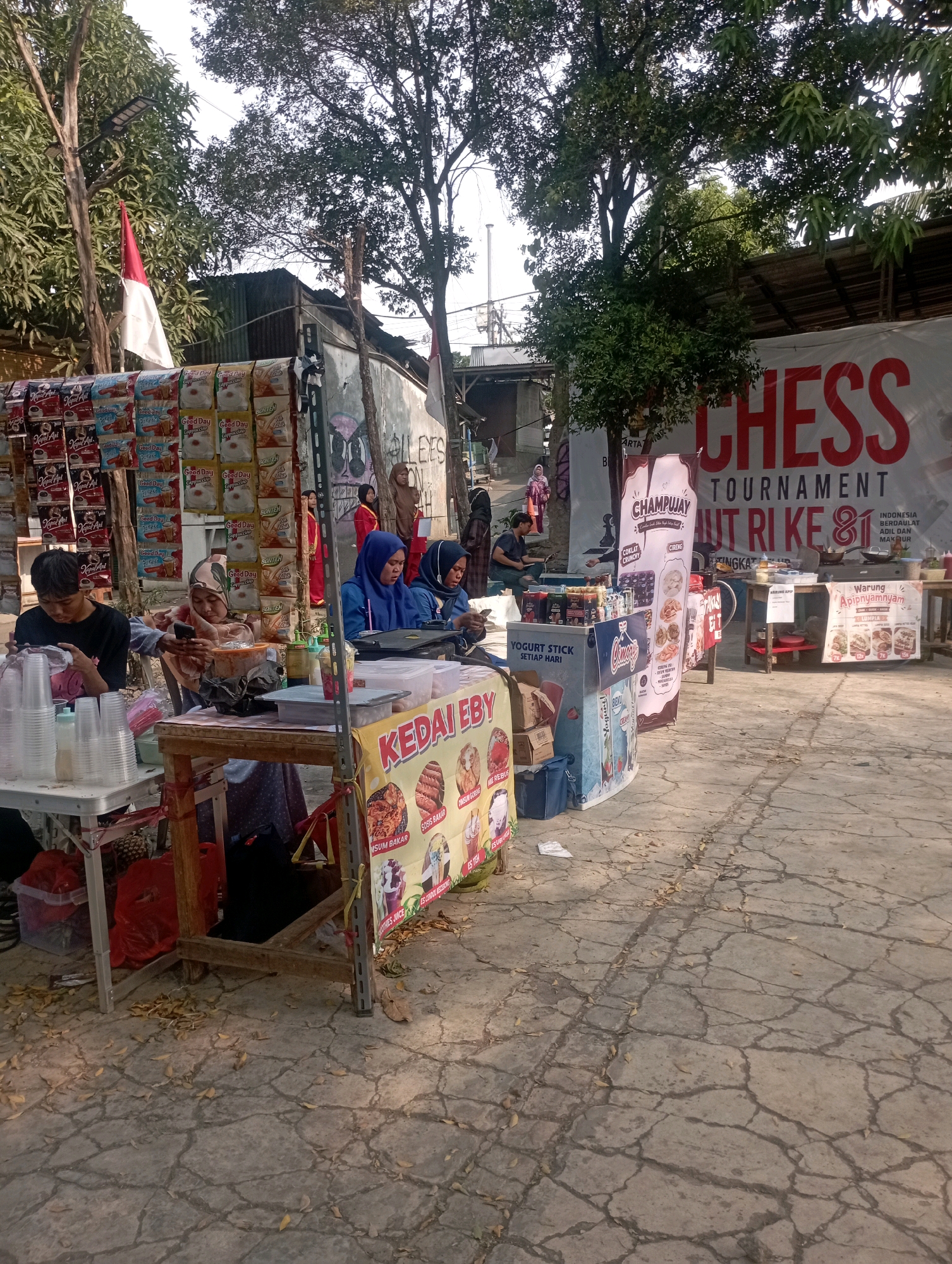 This is a medium shot, taken outdoors, of an informal marketplace in East Jakarta, Indonesia, likely during the day given the lighting. The scene is dominated by several market stalls set up along a cracked concrete path. In the foreground, a table with clear plastic cups and bottles is visible, with a young man in a black shirt sitting behind it, appearing to be working or interacting with his phone. To the right of this table is a stall with a yellow banner that reads "KEDAI EBY," featuring images of food items. Behind this, a woman in a blue hijab and another in a patterned hijab are seated, one appearing to use a phone and the other looking at something in front of her.
Further back, a large white banner with red lettering announces "CHESS TOURNAMENT" and other text, suggesting an event is taking place. To the left of this banner, another stall displays numerous packaged food items, possibly instant coffee or snacks, hanging from the wall. An Indonesian flag, red and white, is visible hanging from a pole. In the background, trees provide shade, and some structures of corrugated metal and wood are present, indicating a somewhat makeshift setup. People in various attire, including hijabs, are milling around or attending to the stalls. The overall impression is of a busy, community-oriented event.