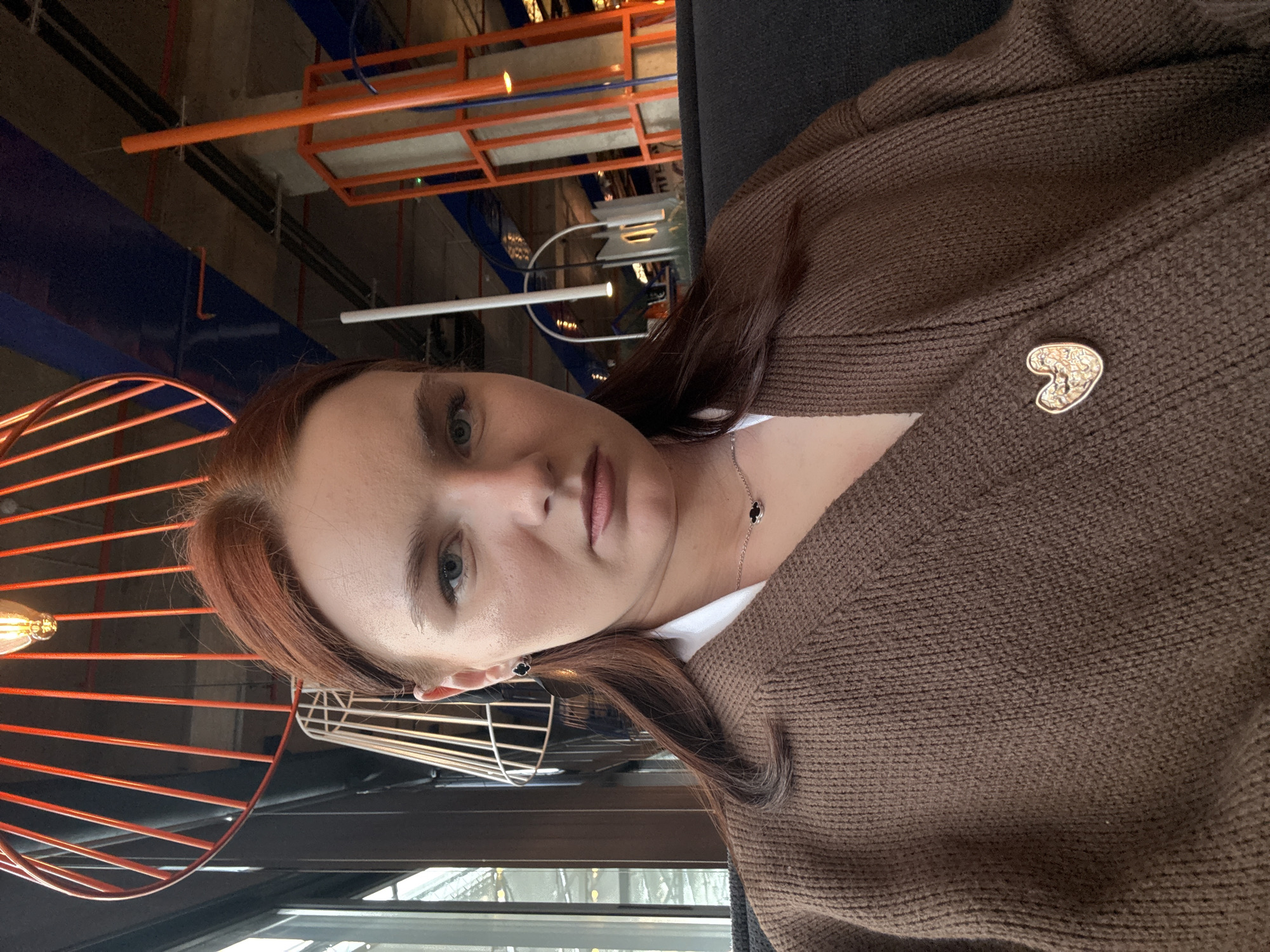 A woman with light skin, auburn hair, and blue eyes is wearing a brown knit cardigan adorned with a heart-shaped pin. She is also wearing a necklace with small black clover-like charms. She is seated near a window, possibly in a café or restaurant. The interior has a modern industrial design, with orange and blue exposed pipes and beams on the ceiling. There is an orange, cage-like light fixture near her. The presence of a window and visible structures suggests a location in an urban setting, possibly Kokkorevo, Russia.