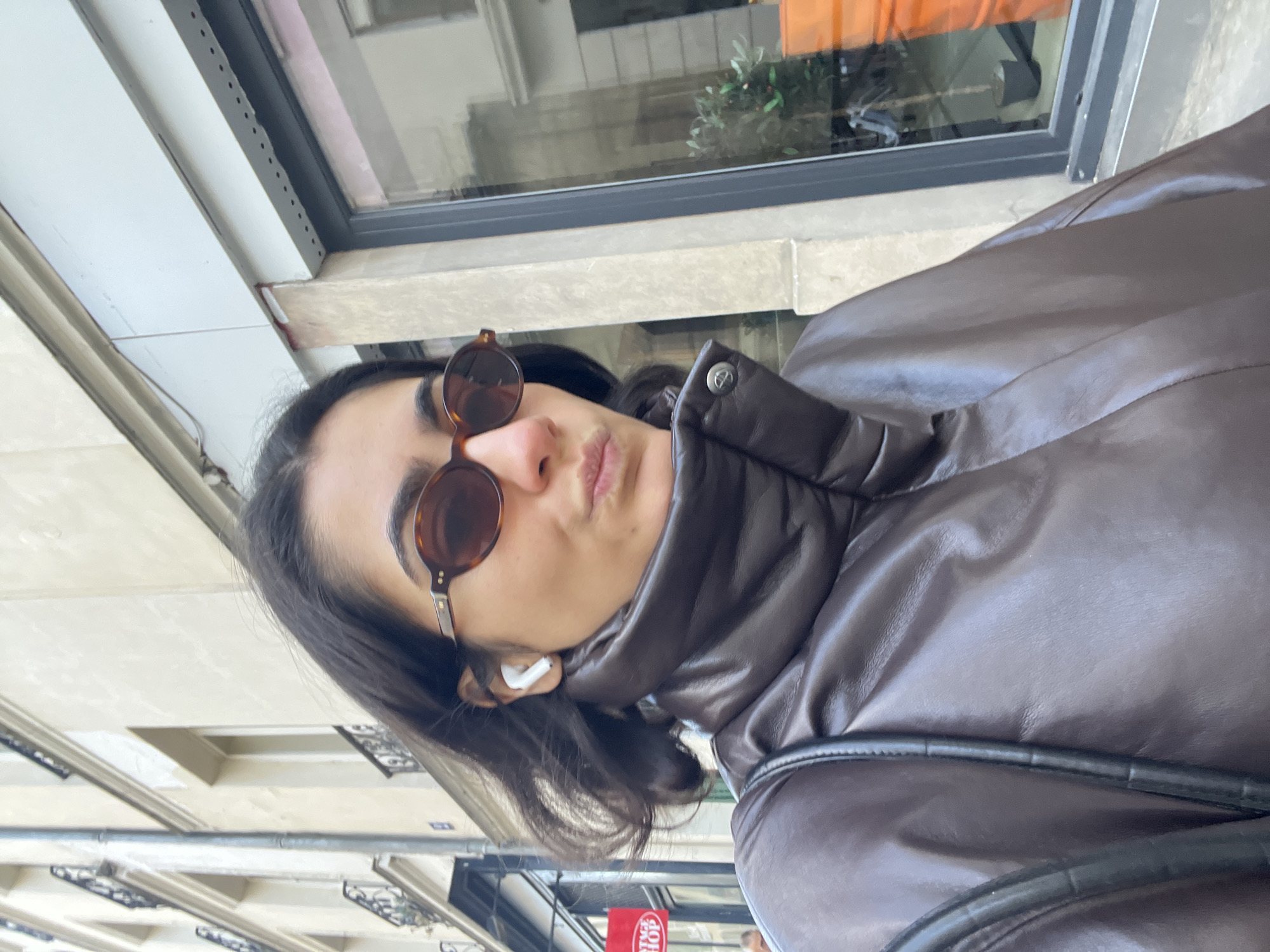 A young person is photographed in an eye-level, medium close-up shot on a city street in Paris, France. The person is wearing a brown, high-necked leather jacket and dark brown sunglasses, with what appears to be a white wireless earpiece visible in their left ear. The individual has dark hair and their lips are slightly pursed.
The backdrop reveals an urban environment: a building's window with a dark frame and glass reflecting an orange-toned object, along with other architectural elements suggesting a Parisian cityscape. A red sign with white text and a portion of a figure are noticeable in the distance. The image is taken during daylight hours.