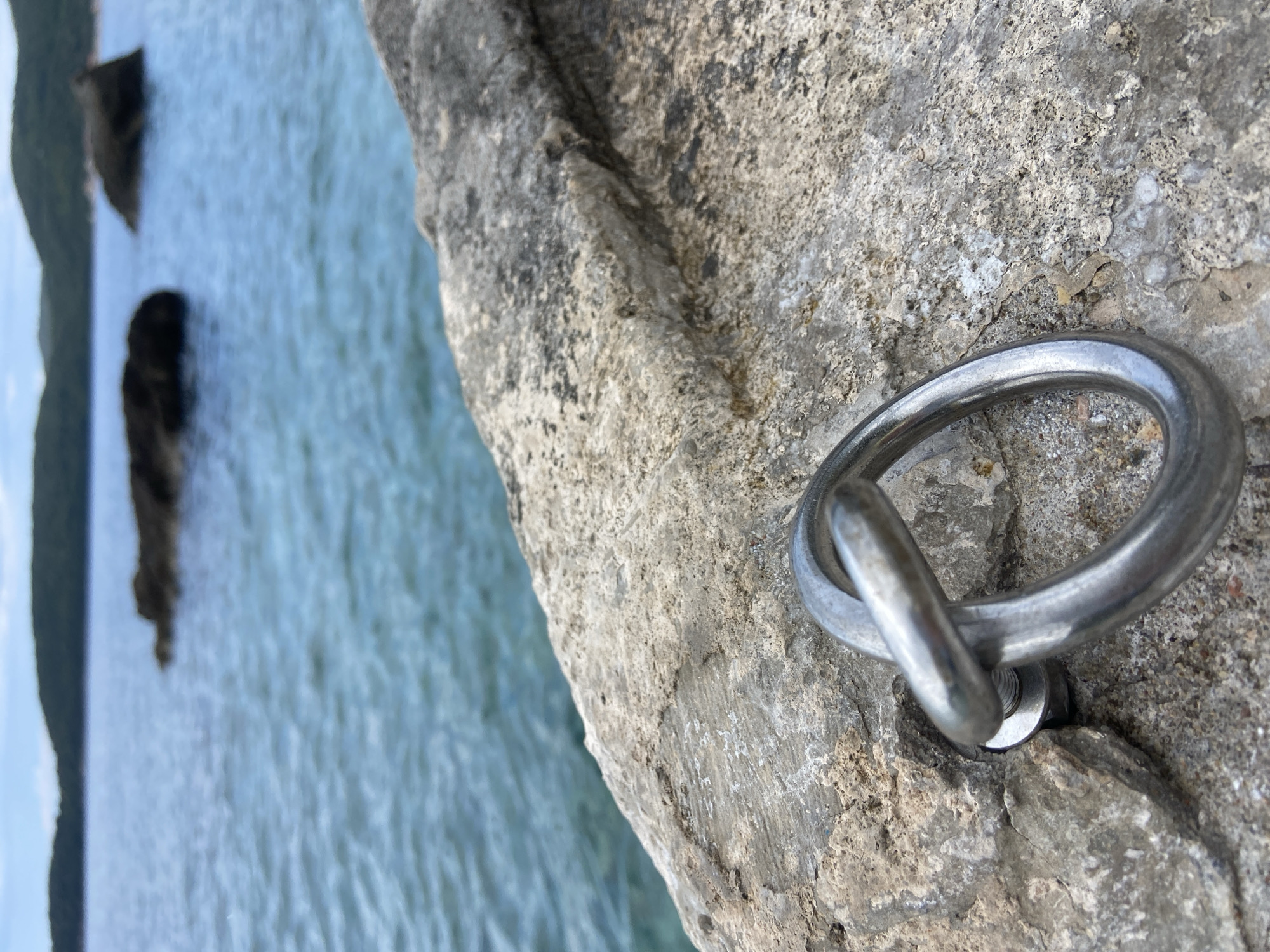 The image shows a metal ring attached to a weathered, gray stone surface. The ring is secured with a bolt and washer. In the background, out of focus, is blue-green water, with dark rock formations visible in the distance. The location is Prijevor, Montenegro.