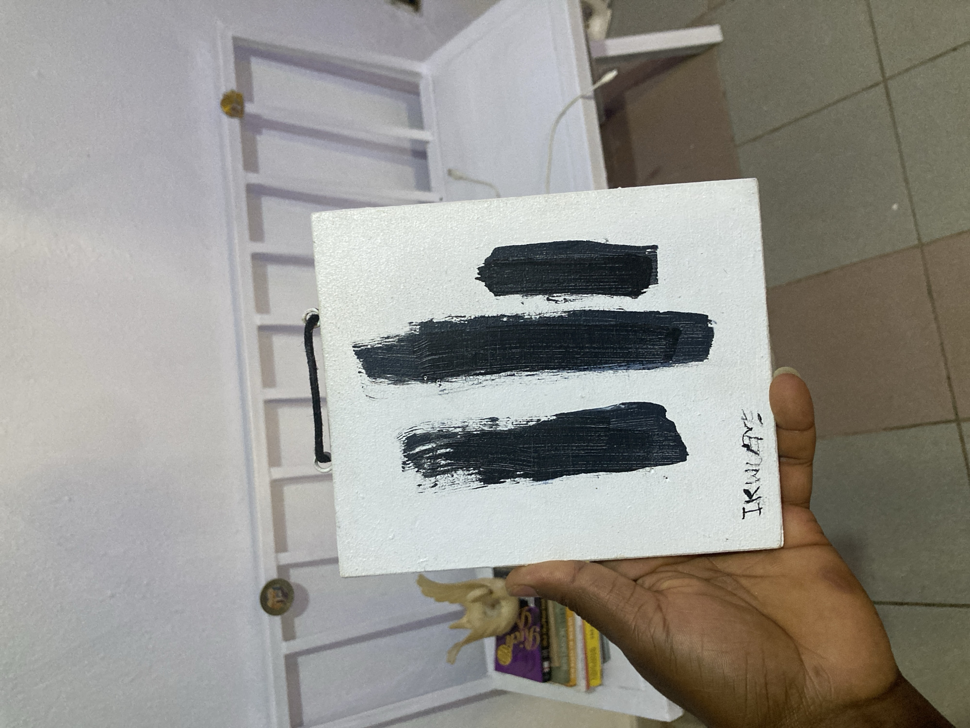 A hand holds a small, white, textured canvas featuring three horizontal black brushstrokes of varying lengths, signed "IKWUOYE" in dark ink in the lower right corner. The background is a white shelving unit with contents including books and a small bird figurine. The photograph may have been taken in a residence in the city of Jos, Nigeria. The floor is covered in light-colored, square tiles.
