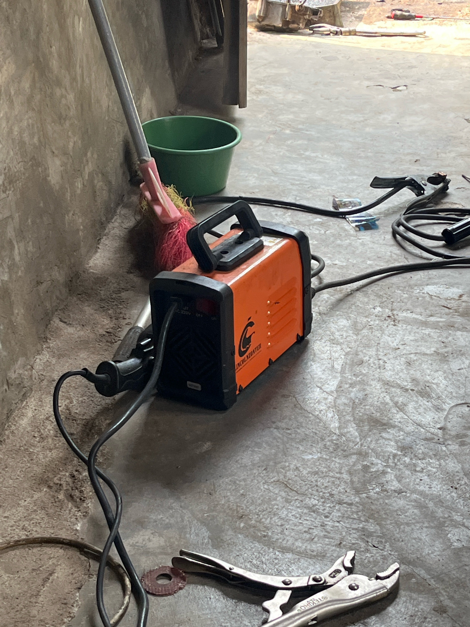 The image depicts a scene that appears to be a workspace or garage, potentially located in Kebumen, Indonesia. The floor is concrete, showing signs of wear and some texture. In the foreground, a pair of locking pliers is visible, resting on the concrete. Adjacent to the pliers is a small, circular, reddish-brown object, possibly an abrasive disc.
Dominating the center of the image is an orange welding machine with a black handle. Black cables extend from the welding machine, one of which is plugged into a black power cord. To the left of the welding machine, leaning against a textured, gray wall, is a mop with a pink head and a silver handle. Next to the mop, a green bucket is situated.
In the background, out of focus, are other objects that suggest a work or storage area, including what appears to be metal machinery and possibly other tools or equipment. The lighting suggests an indoor environment, perhaps with natural light filtering in from an opening. No people are present in the image.