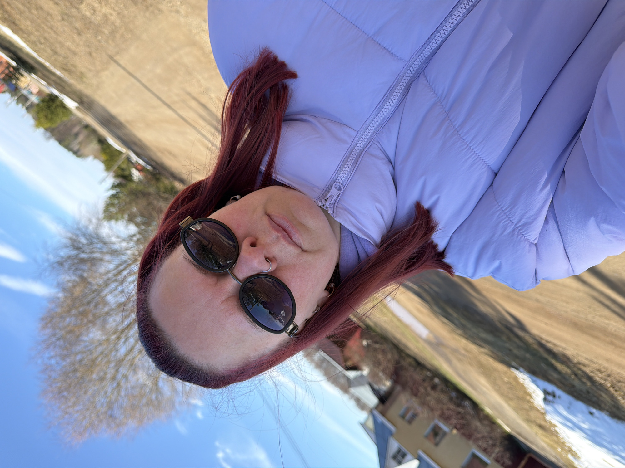 A woman with auburn hair and sunglasses is photographed from a low angle. She wears a light purple puffy jacket. In the background, there is a street with some snow on the ground and buildings. The location is possibly Chaikovsky, Russia.