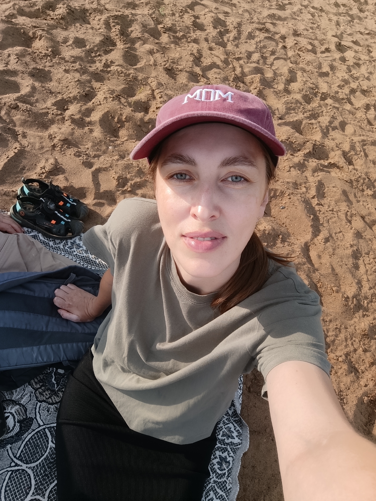 This is a selfie of a woman sitting on a sandy beach, likely in Chaikovsky, Russia. She is wearing a maroon baseball cap with "MOM" embroidered in white letters on the front, a plain olive-green t-shirt, and a black skirt. The woman has light skin, blue eyes, and light brown hair tied back in a ponytail. She is smiling slightly as she looks at the camera.
Beside her, on the sand, are a pair of black and teal sandals. She is sitting on a patterned blanket, and next to that is a dark gray backpack. The sand appears dry and textured, with footprints visible. The lighting suggests it is daytime, likely late afternoon due to the warm glow on her face. The overall impression is one of a relaxed day at the beach.