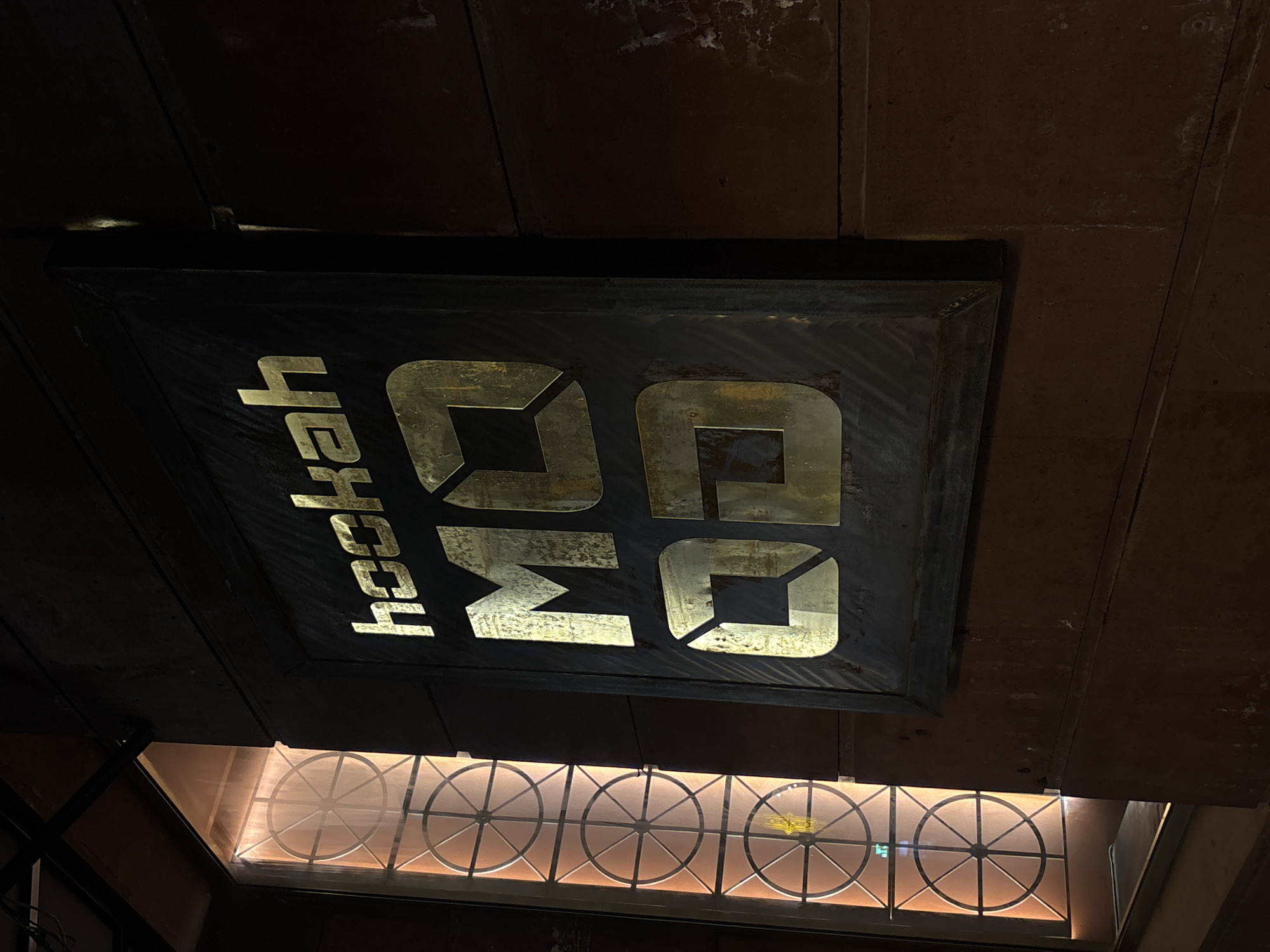 A low-angle shot shows a black sign with gold lettering spelling "hookah" vertically and the letters "OQ" horizontally. The sign hangs from a dark brown or maroon ceiling. Below the sign is a window with a decorative metal grid pattern of circles with radiating lines, lit from behind with a warm glow. Based on visual elements, the location may be Gyumri, Armenia.