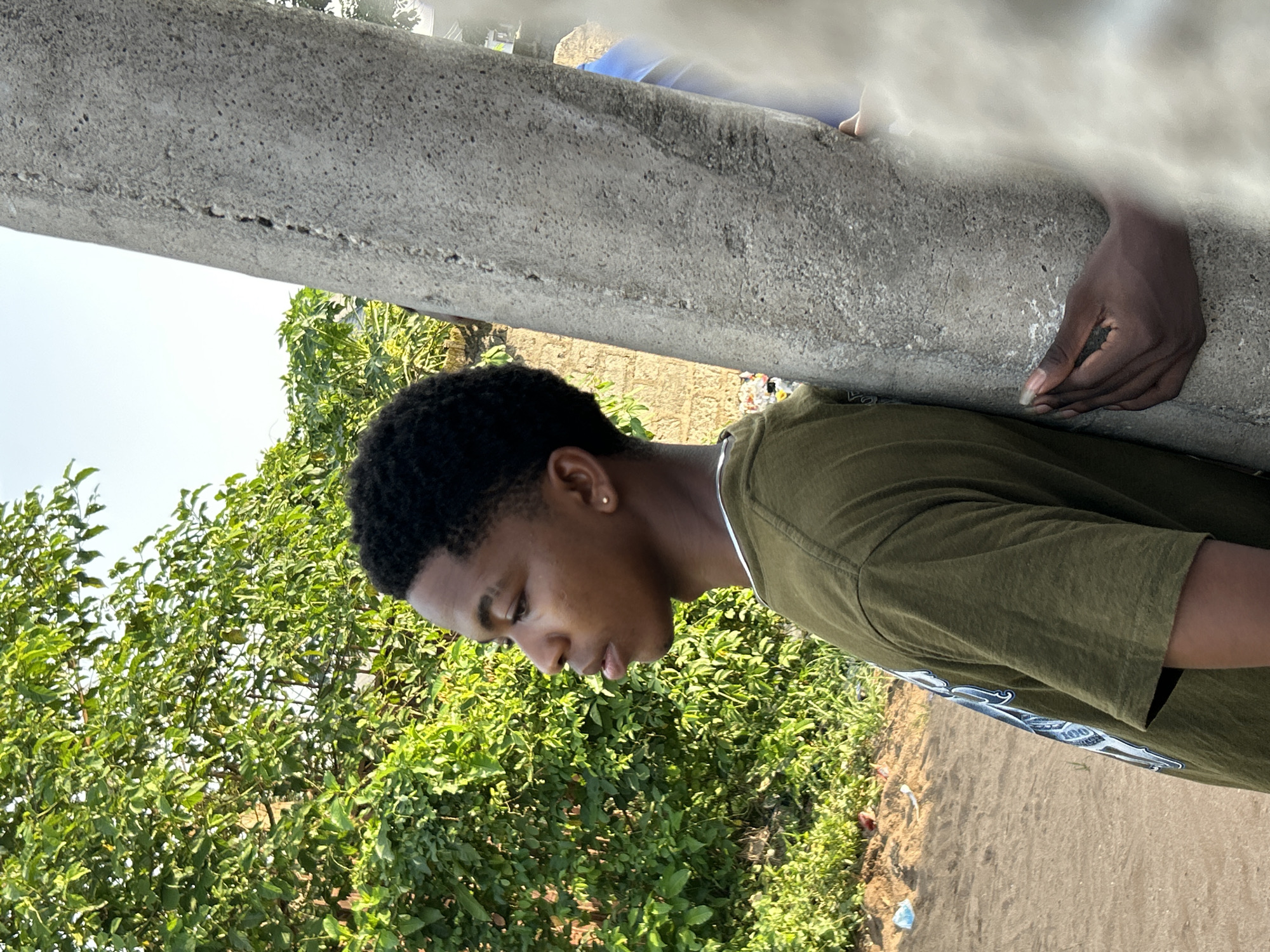 A young, dark-skinned male stands next to a concrete structure in an outdoor setting, likely in Ihiagwa, Nigeria. The individual has short, dark hair and is wearing a green t-shirt with a graphic design. He is looking downward, with his face partially obscured. A hand rests on the concrete structure. Dense green vegetation fills the left side of the frame. The ground is visible on the right side, alongside a paved surface and some debris. The concrete structure is textured, showing signs of weathering. Above the concrete structure, there is a blurry figure.