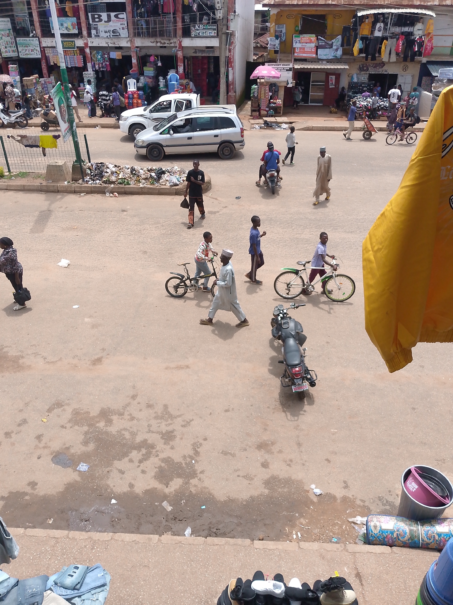 An aerial, high-angle, medium shot captures a street scene in an urban environment, likely a market area, in front of shops. In the foreground, a person wearing a yellow jacket is partially visible on the right, obscuring the view. The street is paved with asphalt and shows signs of wear and tear, with some damp patches and scattered litter, including plastic bags and debris.
Several people are present in the scene, most of them walking or interacting with vehicles. On the street, a black motorcycle is parked, and another person is riding a bicycle. Further back, two cars are visible: a white SUV and a silver minivan. In the background, a multi-story building houses various shops, with awnings and merchandise displayed. The overall impression is one of a busy, commercial street with a mix of pedestrian and vehicular traffic.