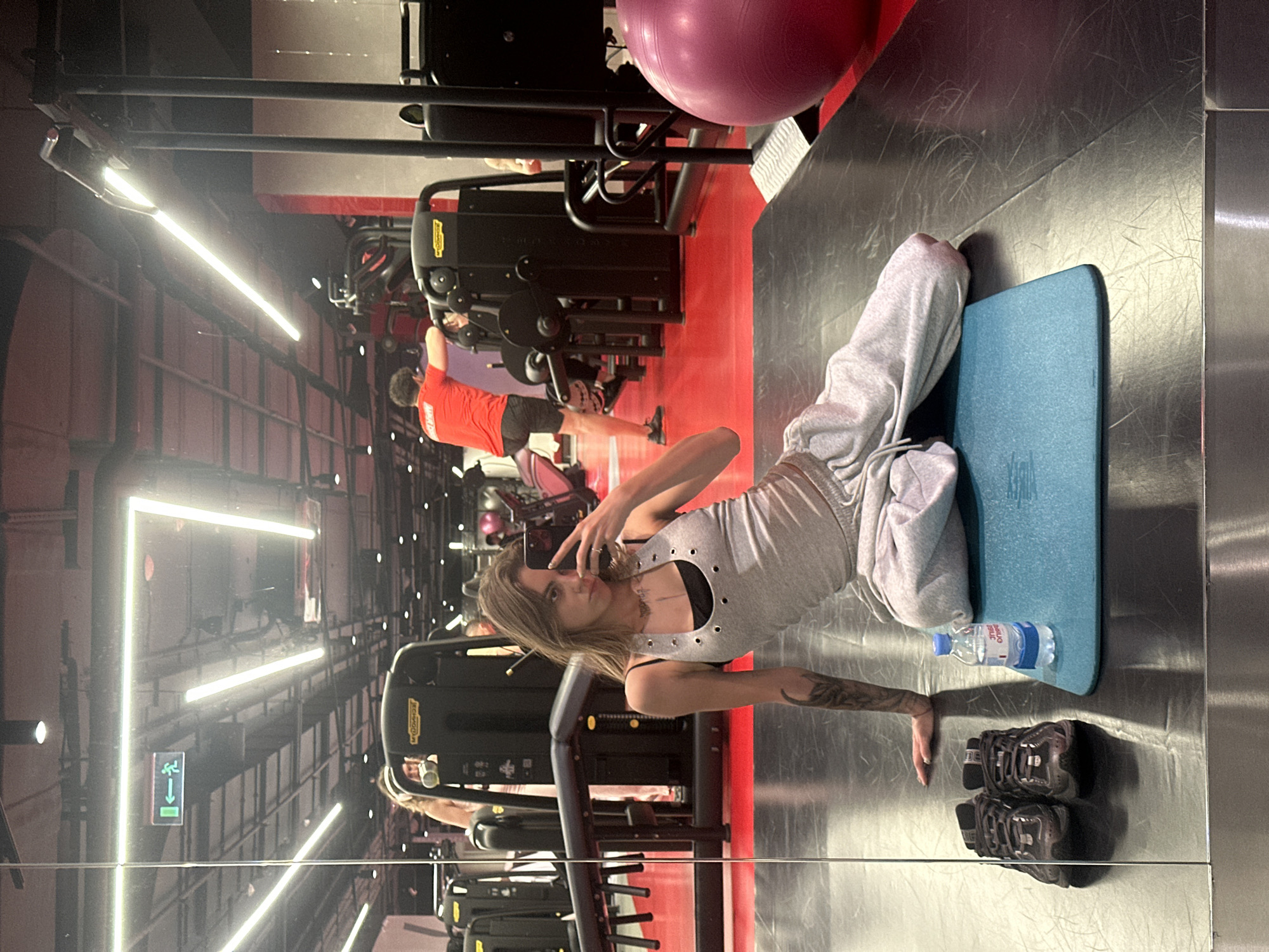 A woman with tattoos on her arm is sitting on a blue yoga mat in a gym. She is wearing a grey tank top and grey sweatpants. She is holding a phone and taking a selfie. There are a pair of black sneakers and a bottle of water next to the mat. In the background, there are gym equipment and other people working out. The gym appears to be modern and well-lit with red and black flooring and ceiling lights.