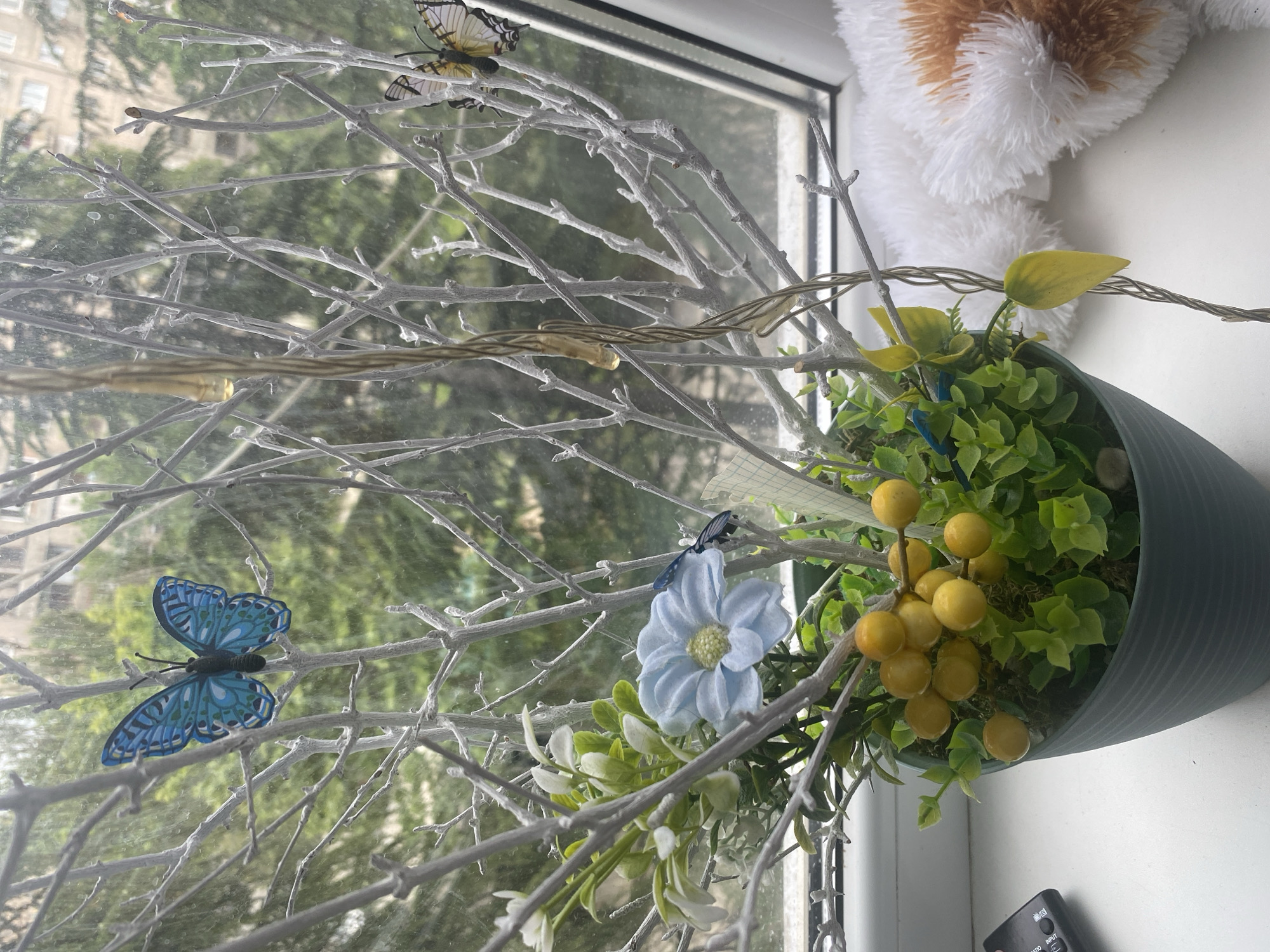 A still life image depicts a potted artificial plant with light green foliage and yellow berry-like decorations, placed on a white windowsill. The plant is adorned with decorative branches painted white, and artificial butterflies in blue and yellow hues. A string of lights with yellow bulbs is also entwined among the branches. In the background, visible through a window, is a blurred view of green trees and a building. To the right of the plant, a plush toy with white and orange fur is partially visible. To the left, a black remote control rests on the windowsill. The scene is illuminated by natural light filtering through the window.