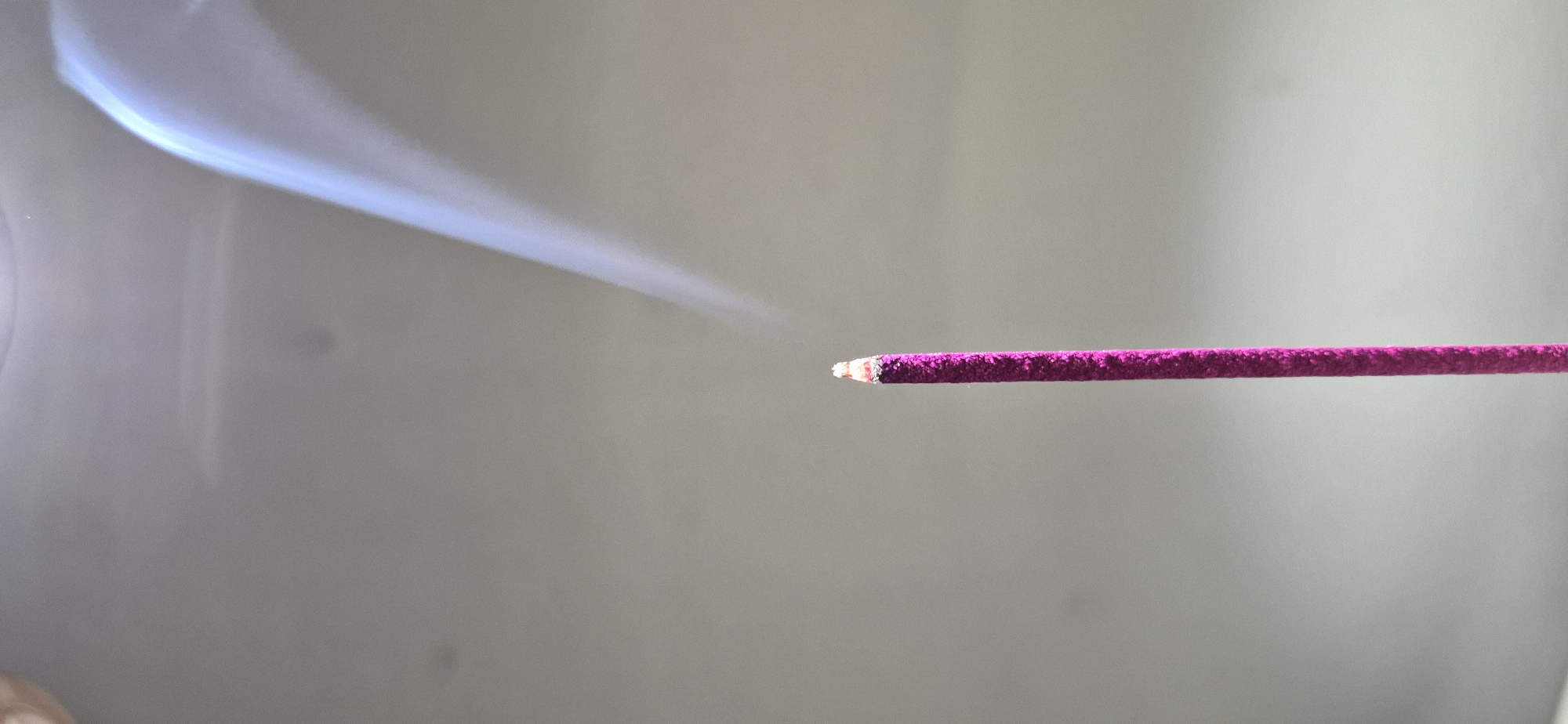 The image shows a close-up of a lit pink incense stick with smoke rising from its tip against a plain, light-colored background. The incense stick is positioned vertically in the center of the frame, with its burning tip glowing red and emitting a thin stream of white smoke that curves upwards and to the right. The background is a soft, out-of-focus gradient of gray, which highlights the incense stick and the smoke. There is no discernible location in the city of Ussurijsk, Russia, visible in this image.