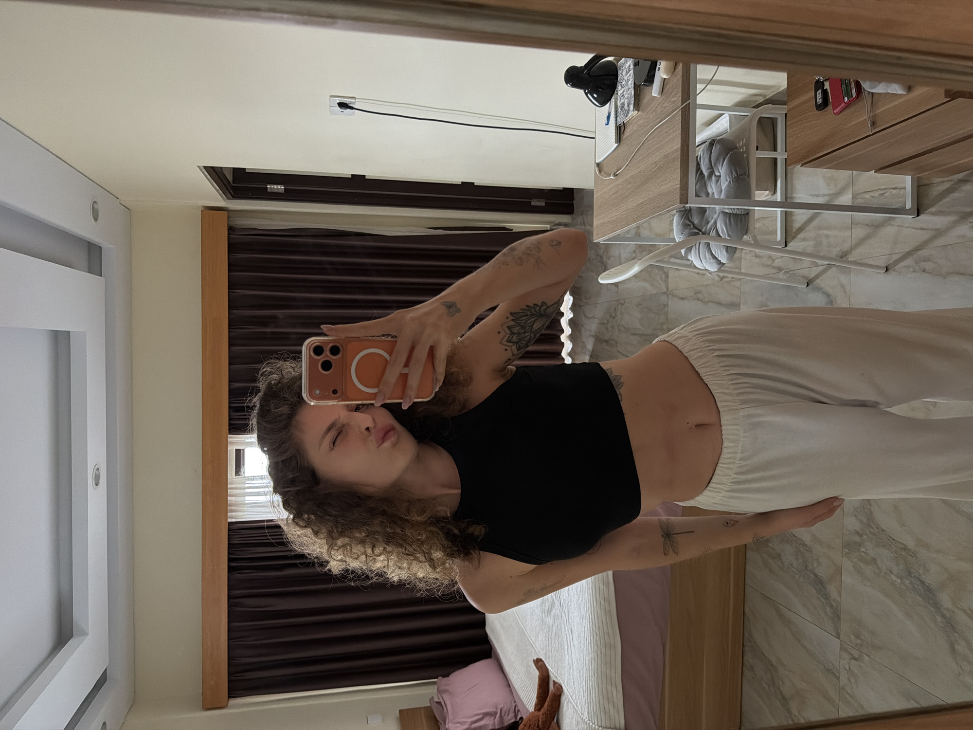 A young woman with curly brown hair, wearing a black crop top and white pants, poses for a selfie in a bedroom. She is winking and puckering her lips. In the background, a bed with pink and white bedding is visible, along with dark brown curtains. A desk with a lamp, notebook, and electrical outlet is also in the room. The floor is tiled with a marble pattern. The scene appears to be in North Kuta, Indonesia.