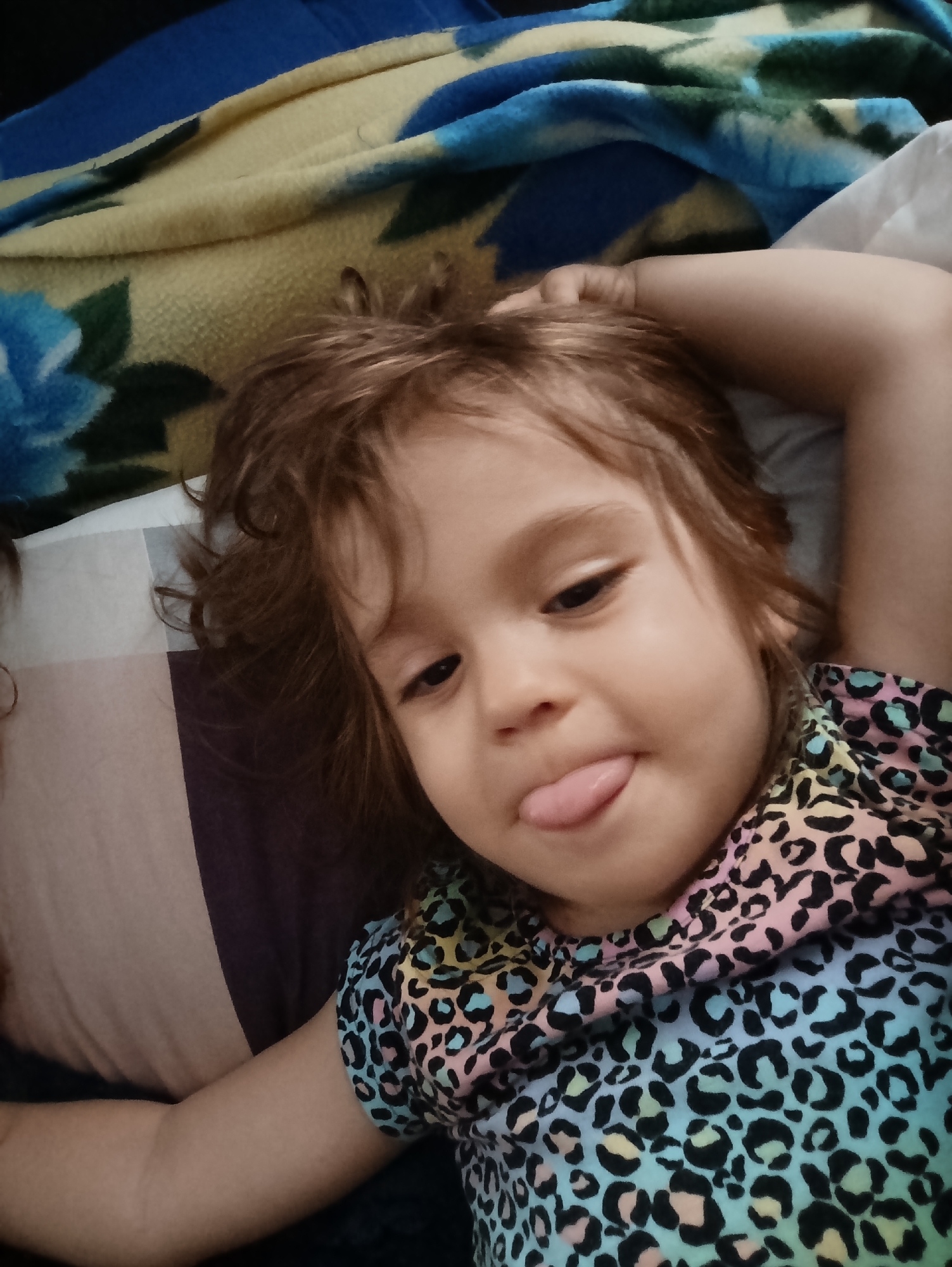 An indoor, close-up photograph captures a young child, likely a girl, lying down and looking directly at the camera with a playful expression, her tongue sticking out. She has light brown, somewhat curly hair and is wearing a vibrant, multi-colored top adorned with a leopard print pattern in shades of blue, pink, and yellow. Her right arm is bent, with her hand resting above her head, suggesting a relaxed posture.
The child is situated on a bed or soft surface. A patterned blanket or comforter with blue floral designs, possibly roses, against a yellow and blue background, is visible beneath her head and upper body. To her left, a striped pillow or blanket in hues of brown, purple, and cream is partially visible, while a white pillow can be seen to her right. The scene conveys a casual and lighthearted moment, possibly during leisure time in a home environment. There are no discernible external light cues to indicate the time of day. The location is Новоалександровка, Russia. No text is visible in the image.