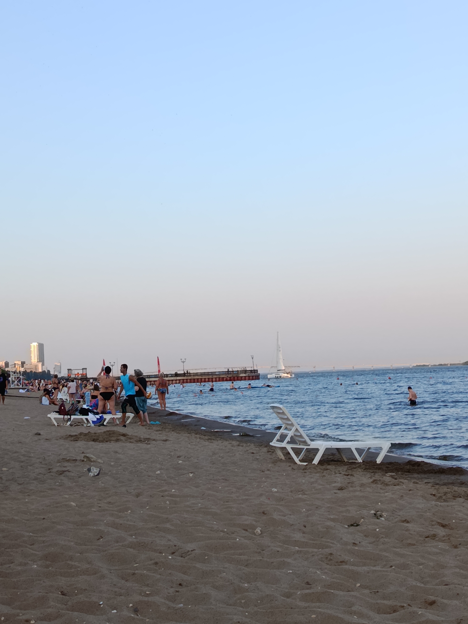 A beach scene in the city of Unknown, Unknown, shows numerous people enjoying the sand and water under a clear, pale sky. In the foreground, a sandy beach is sparsely populated with a few white lounge chairs. Further back, a cluster of people are gathered, some standing and interacting, while others are seated or walking. The water is a vibrant blue, with several individuals swimming and wading. A pier extends into the water, with more people visible along its length. A white sailboat is moored in the distance, and a bridge can be seen in the far background. The overall atmosphere is relaxed and recreational, with a sense of a warm, sunny day at the beach.