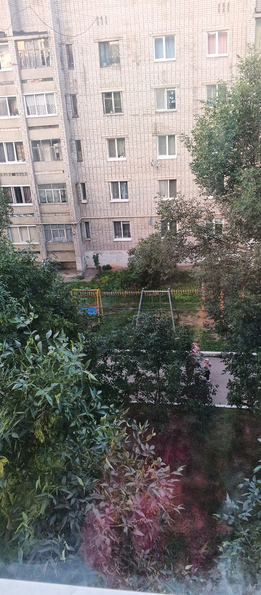 This is a high-angle, full-frame, vertical shot of a residential courtyard in Izhevsk, Russia, likely taken from a window of a multi-story apartment building. The building, constructed of light-colored brick, dominates the upper portion of the image. It features numerous windows, some with balconies, and appears to be an older Soviet-era panel building.
Below the building, a children's playground is visible, with a swing set. The courtyard area is green, with grass and trees, indicating a relatively temperate climate. Two individuals, appearing to be women or older girls, are walking on a paved path in the lower right quadrant of the image, partially obscured by dense foliage from trees in the foreground. The overall impression is a typical urban residential scene, with elements of both built environment and nature.