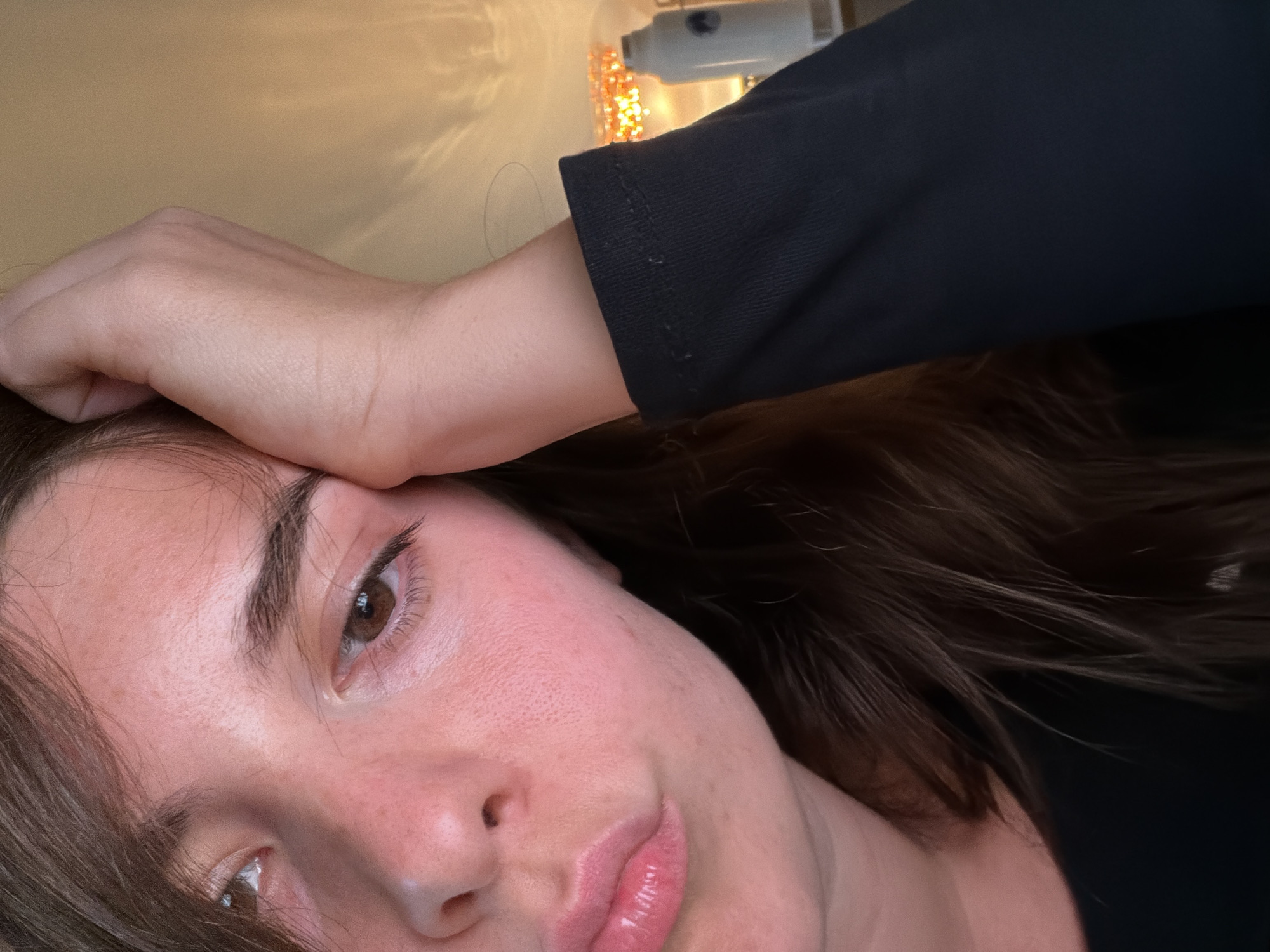 A close-up, slightly tilted, indoor shot shows a woman with light skin, brown hair, and brown eyes. She is wearing a black long-sleeved shirt and has her right hand resting on her forehead. The background includes a blurred light source and a beige wall. The location cannot be determined based on the image.