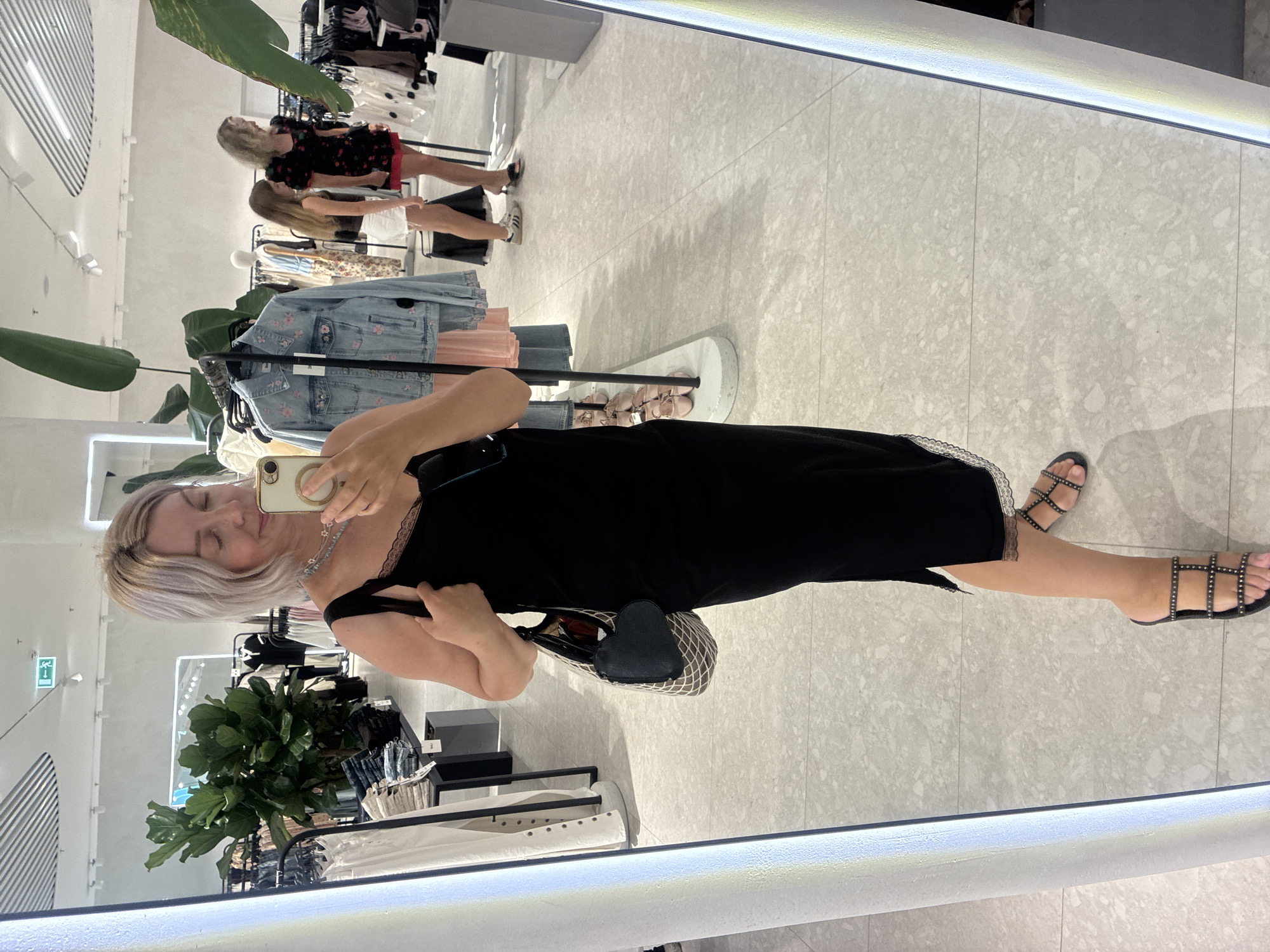 A woman is taking a mirror selfie in a clothing store. She is wearing a black slip dress with lace trim and black studded sandals. She is holding a beige smartphone in her right hand, and her left hand is resting on her purse, which is a beige mesh tote bag with a black heart-shaped coin purse attached. In the background, racks of clothing are visible, as well as other shoppers. The store appears to be well-lit and modern, with tiled floors and a white ceiling. The woman's hair is blonde and styled in a bob. She is looking at her reflection with a slight smile. The location is not specified as Sochi, Russia.