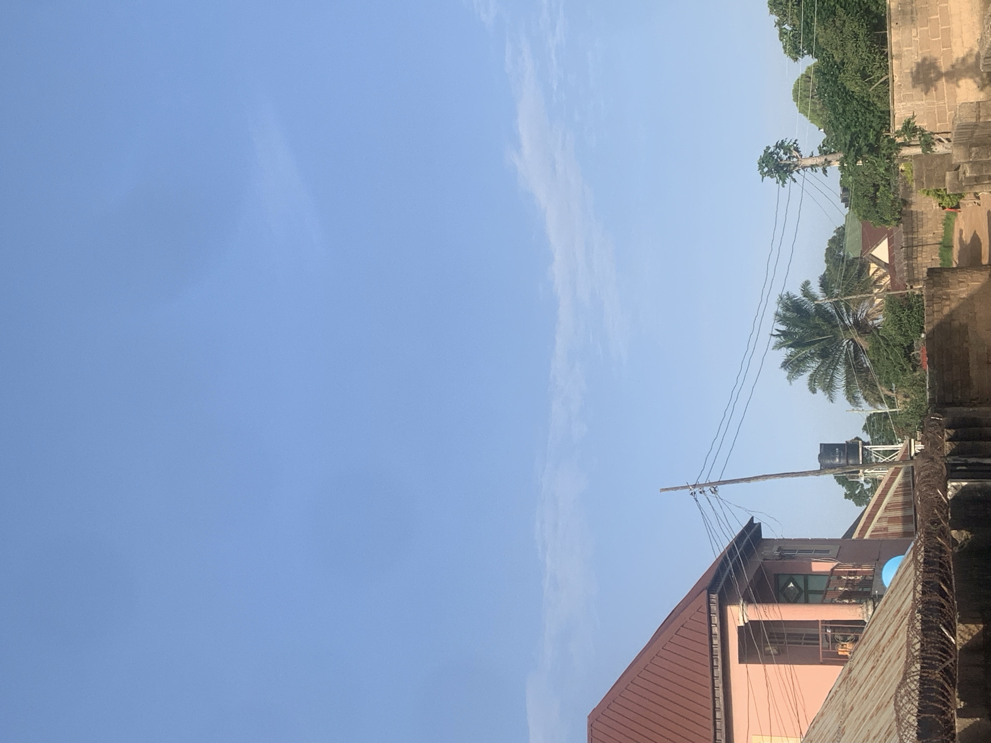 The image depicts a residential area in Jos, Nigeria, under a clear blue sky with scattered wispy clouds. In the foreground, a portion of a pink building with a rust-colored metal roof is visible on the left, featuring a balcony with a railing. Adjacent to it is a corrugated metal roof, partially obscured by what appears to be barbed wire fencing. A wooden utility pole with multiple power lines runs vertically through the scene. In the background, a black water tank on a stand is noticeable. Lush green trees, including a palm tree, dot the landscape, interspersed with other buildings and brick walls. The overall impression is that of a typical urban or semi-urban setting in Jos.