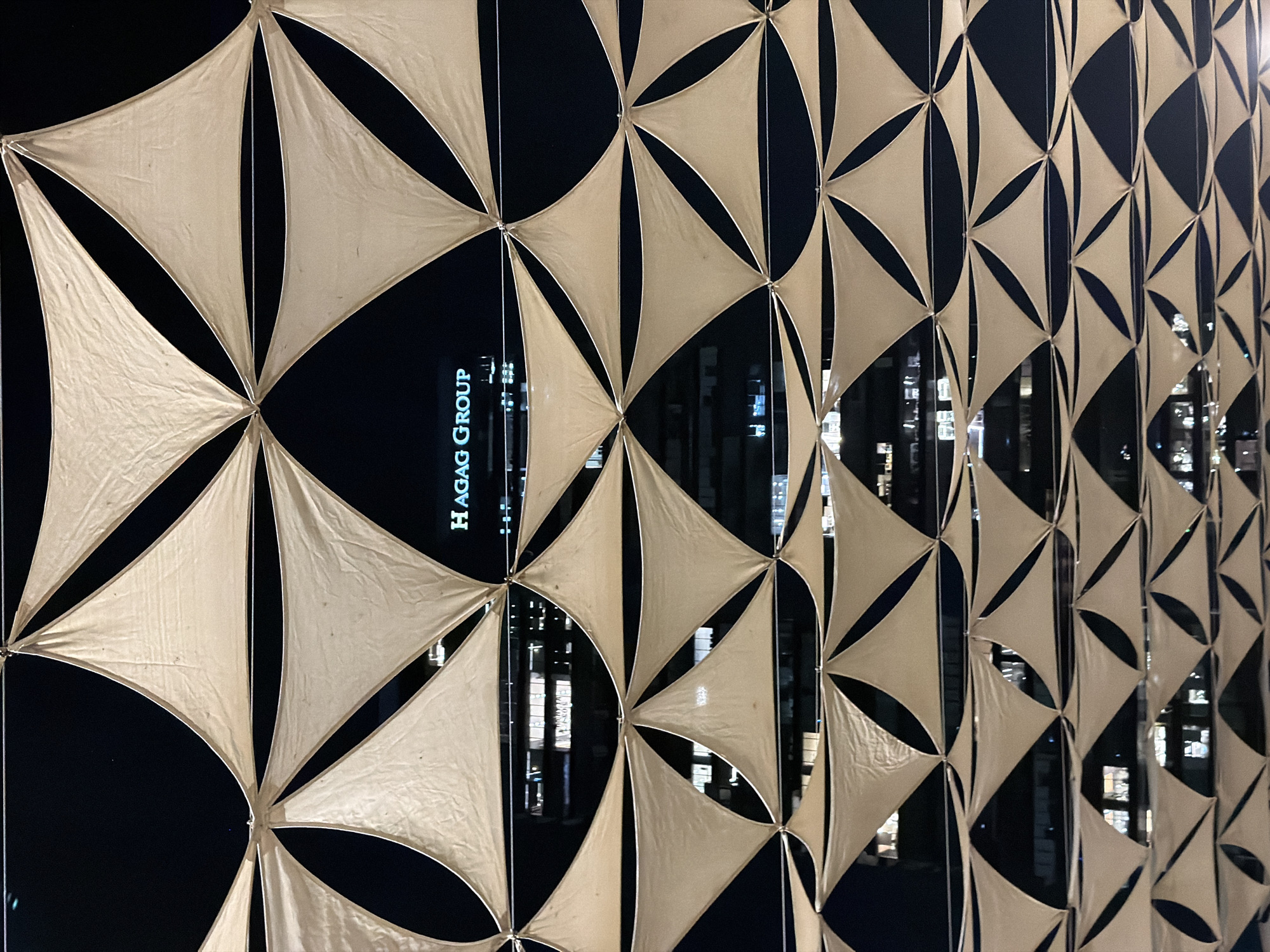 The image shows a nighttime view of a building facade partially obscured by a canopy of beige triangular fabric sails. The sails are stretched taut and arranged in a repeating pattern, creating diamond-shaped openings. Behind the sails, a modern building with illuminated windows is visible. The building has a prominent sign that reads "HAGAG GROUP" in white illuminated lettering against a dark background. The overall impression is of an urban environment at night, likely a public space or a commercial area. The image appears to be taken from a low angle, looking upwards at the canopy and building. The specific location in Tel-Aviv, Israel is not discernible from the image itself, but the "HAGAG GROUP" sign suggests a commercial or residential development by that company, which is known to operate in Israel.