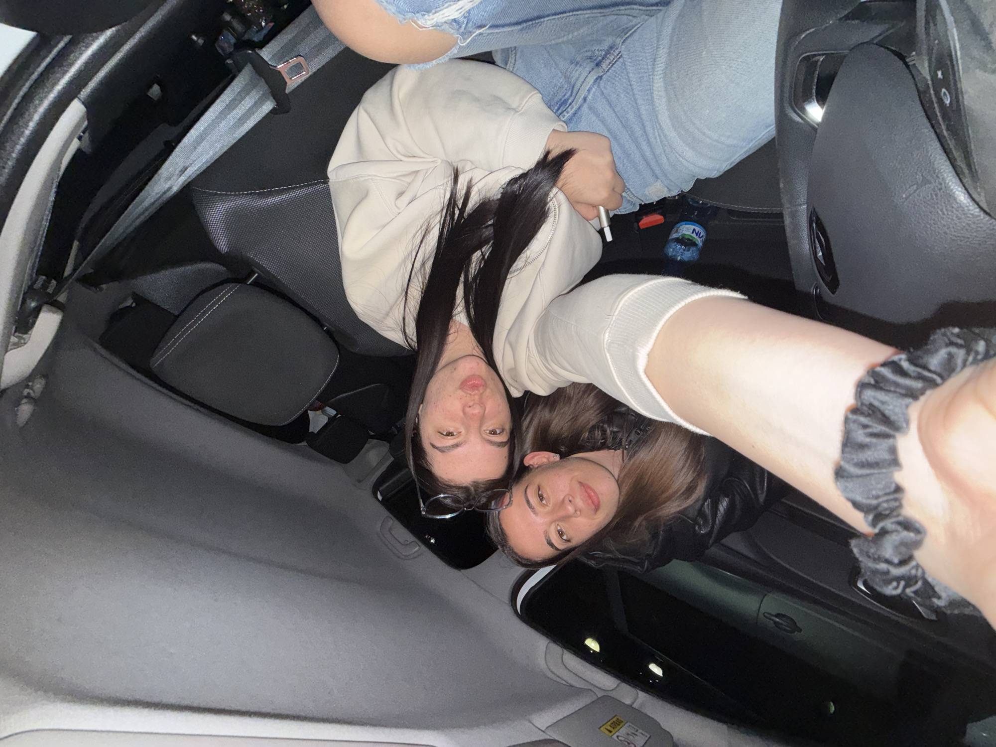 Two individuals are inside a vehicle, possibly a car, with a light gray interior. The car is outfitted with a black seat, a gray seat belt, and a clear bottle of water positioned to the right of the seat. One individual is wearing a beige hoodie and blue jeans, while the other appears to be wearing a black jacket. The photograph appears to have been taken from the passenger seat, capturing both individuals looking at the camera. Location within the city of Karnobat, Bulgaria, cannot be ascertained from this image.