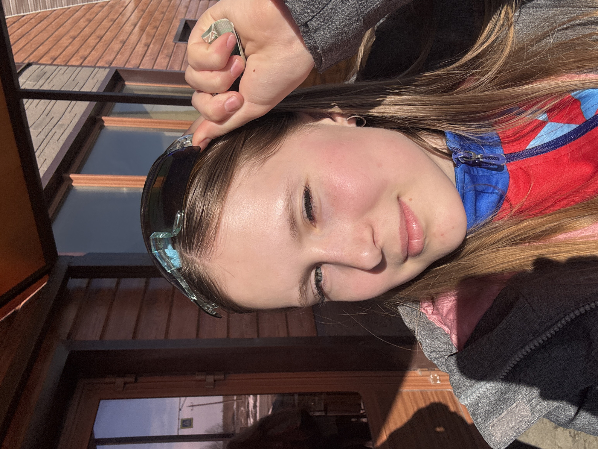 A close-up portrait of a young woman with long brown hair is taken outdoors in Gory, Russia. She is wearing ski goggles pushed up on her forehead and a multi-layered jacket consisting of pink, red, and blue sections, topped with a gray outer layer. Her hand is resting on her forehead, holding a small, square object. The background features wooden walls with windows and the edge of a roof. The image is rotated sideways.