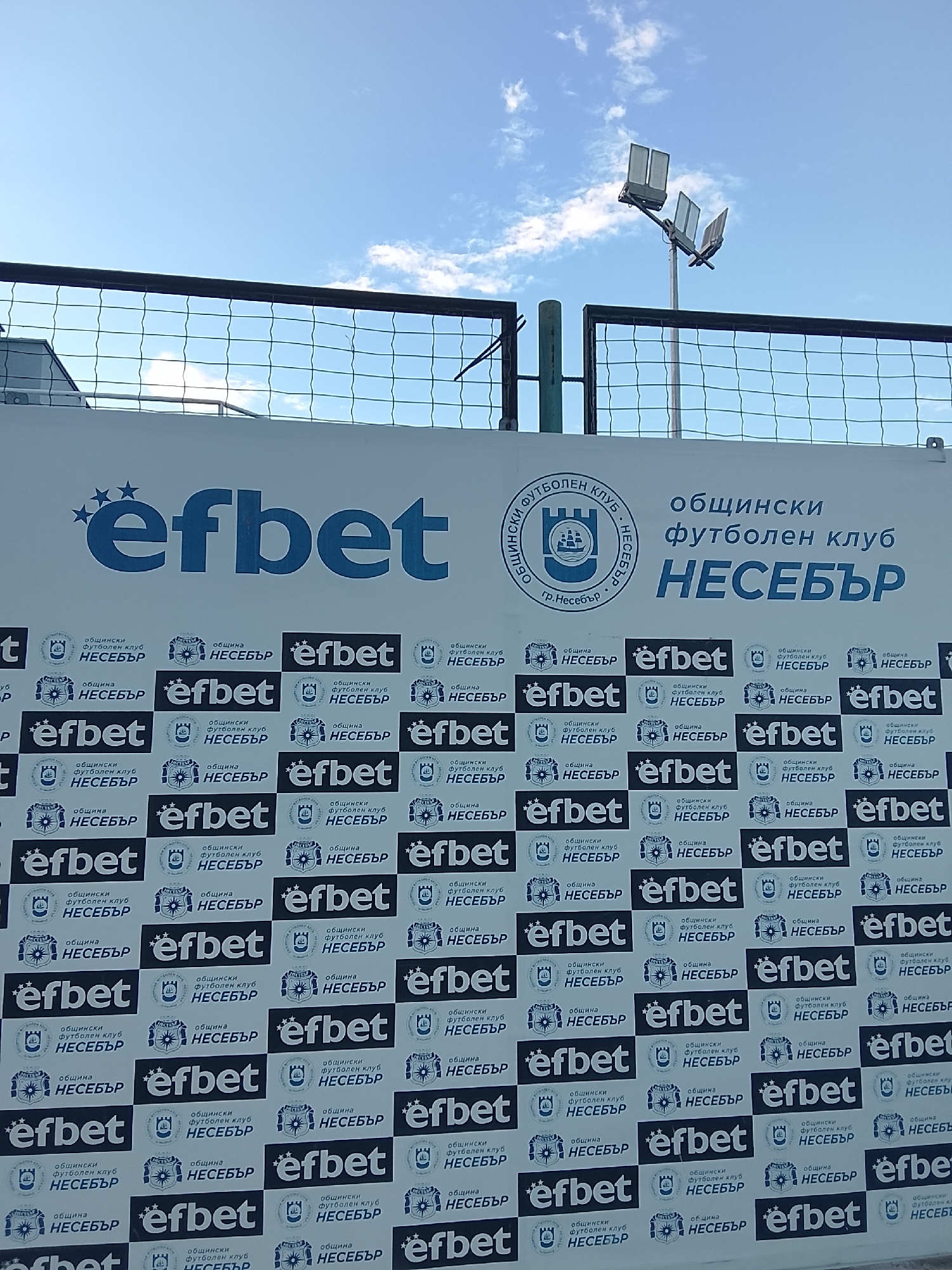 The image displays a large banner with repeated logos and text related to "efbet" and "общински футболен клуб НЕСЕБЪР" (Municipal Football Club Nesebar). The banner is set against a sky with scattered clouds and some fencing and stadium lighting are visible in the upper portion of the image. The banner is likely located at or near the football stadium in Nesebar, Bulgaria, serving as advertisement or sponsorship display. No people are present in the frame.