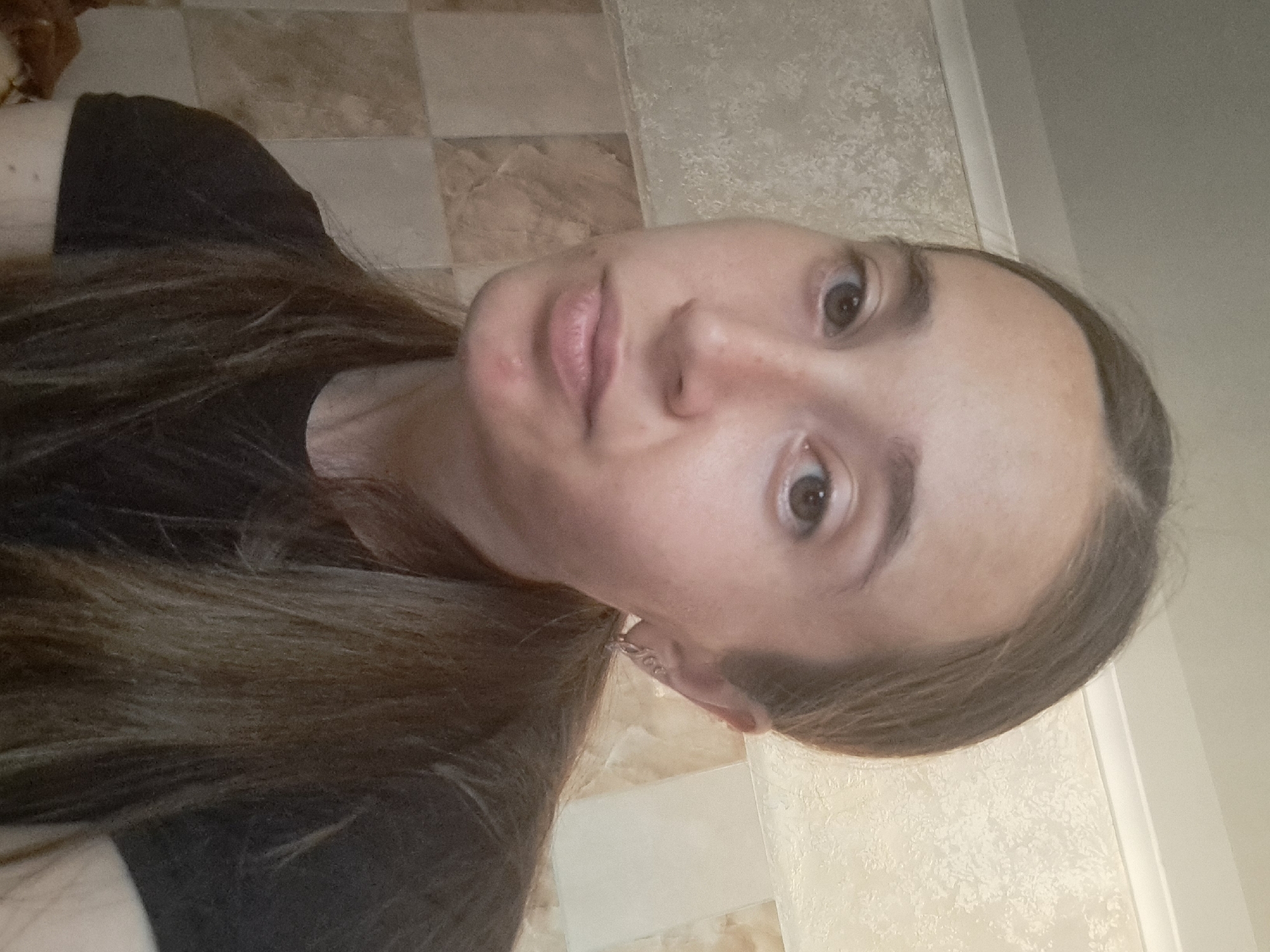 The image is a medium shot of a young woman with long, brown hair tied in a ponytail. She is wearing a black t-shirt and has a small earring in her left ear. Her skin appears to have some blemishes on her chin. The background consists of tiled walls, one with a checkered pattern of beige and brown tiles, and the other with a textured, light-colored finish. The lighting suggests an indoor setting, possibly a bathroom due to the tiling. There are no visible signs of the city of Vypasne, Ukraine in the image.