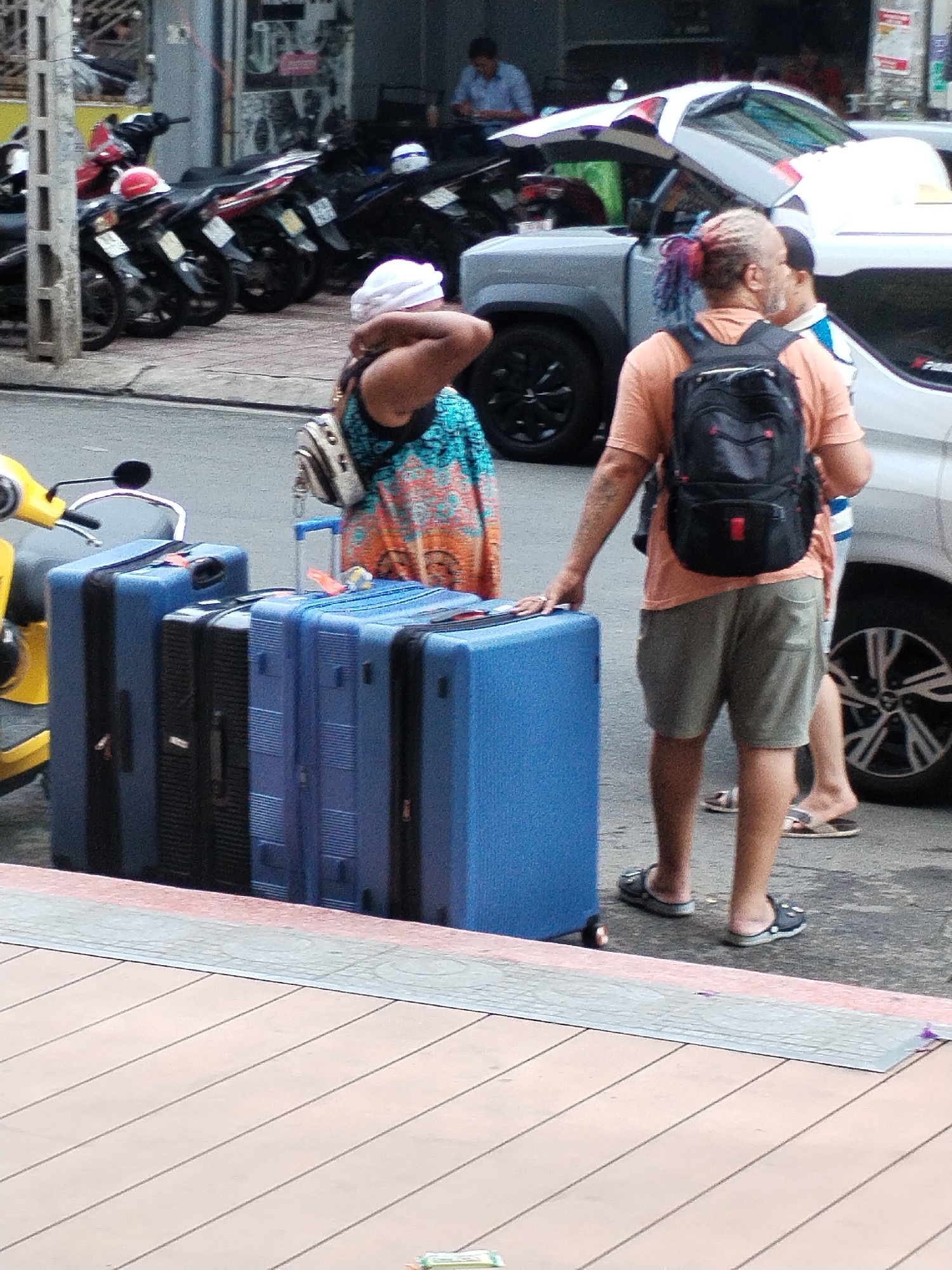 A street in Bac Nha Trang Ward, Vietnam, features three people with several pieces of luggage near a white SUV. A woman with a white head covering and a patterned dress is on the left, standing next to three large suitcases: two blue and one black. A man with multicolored hair and a backpack stands next to her, touching one of the suitcases. A third man is partially visible to the right, wearing a striped shirt and sandals. A row of parked motorbikes lines the street behind them, and a man sits inside a building in the background.