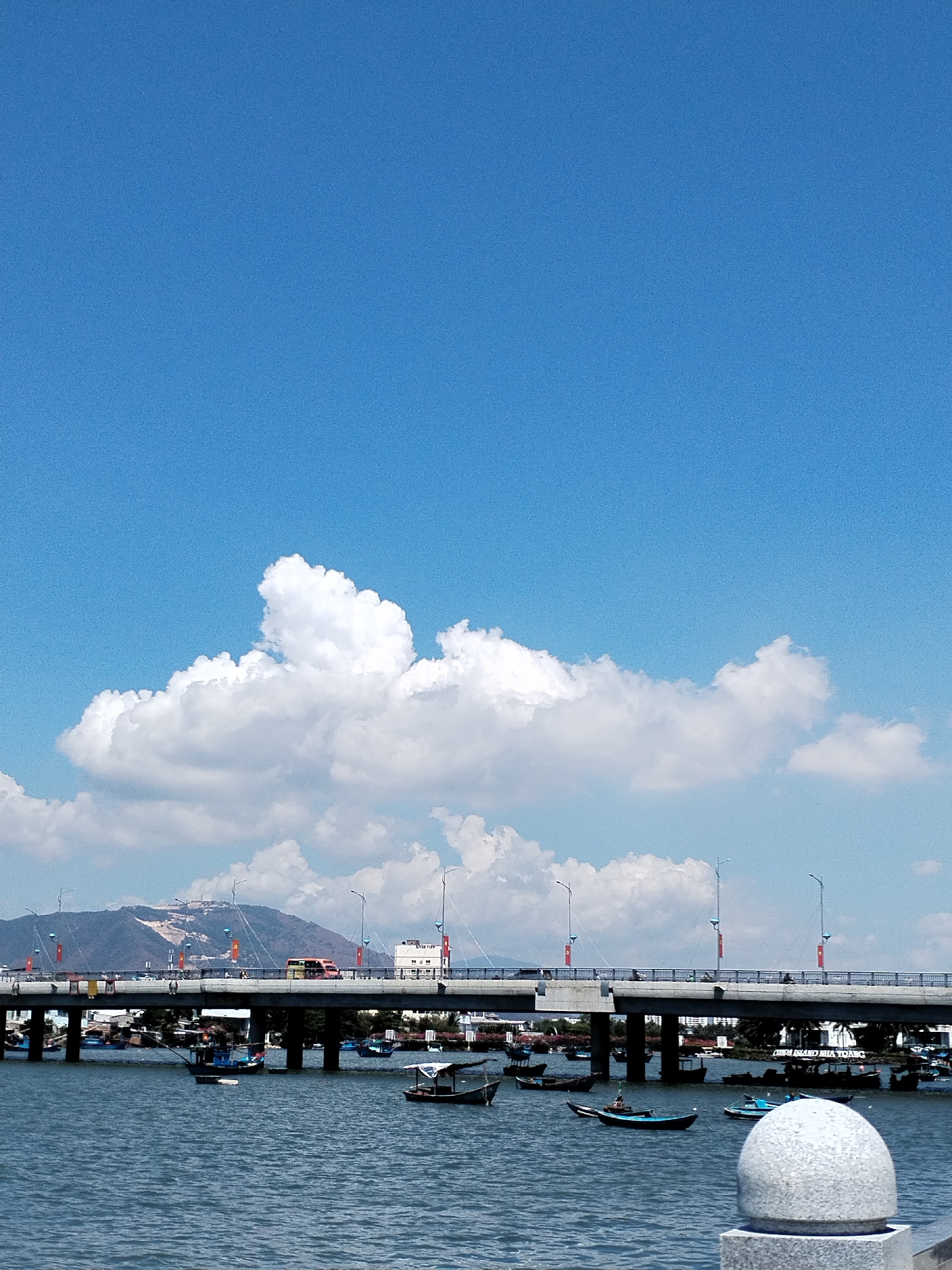 The image depicts a bridge spanning a body of water, likely a river or bay, in what appears to be the Bac Nha Trang Ward of Nha Trang, Vietnam. The bridge is a modern structure with multiple lanes of traffic, visible vehicles including buses, and streetlights. Numerous small boats are scattered on the water beneath and around the bridge. In the background, a mountain range is visible under a partly cloudy blue sky. In the foreground, a concrete post with a spherical top is visible.
