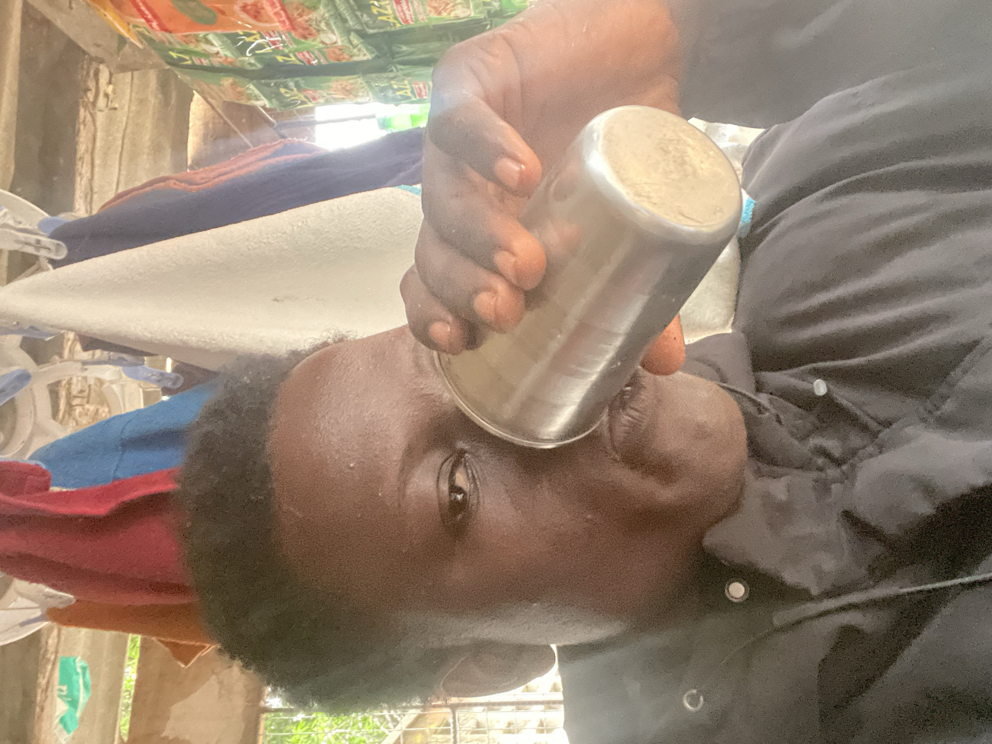 A person with dark skin and short, dark hair is drinking from a metal cup. The person is wearing a dark-colored jacket. Clothes are hanging to dry on a line in the background, secured by clothespins. In the far background, outside the building, green vegetation is visible, suggesting an outdoor environment. The photo appears to be taken in an informal setting, possibly a residential area. I do not have enough information to determine if this photo was taken in Badagry, Nigeria.