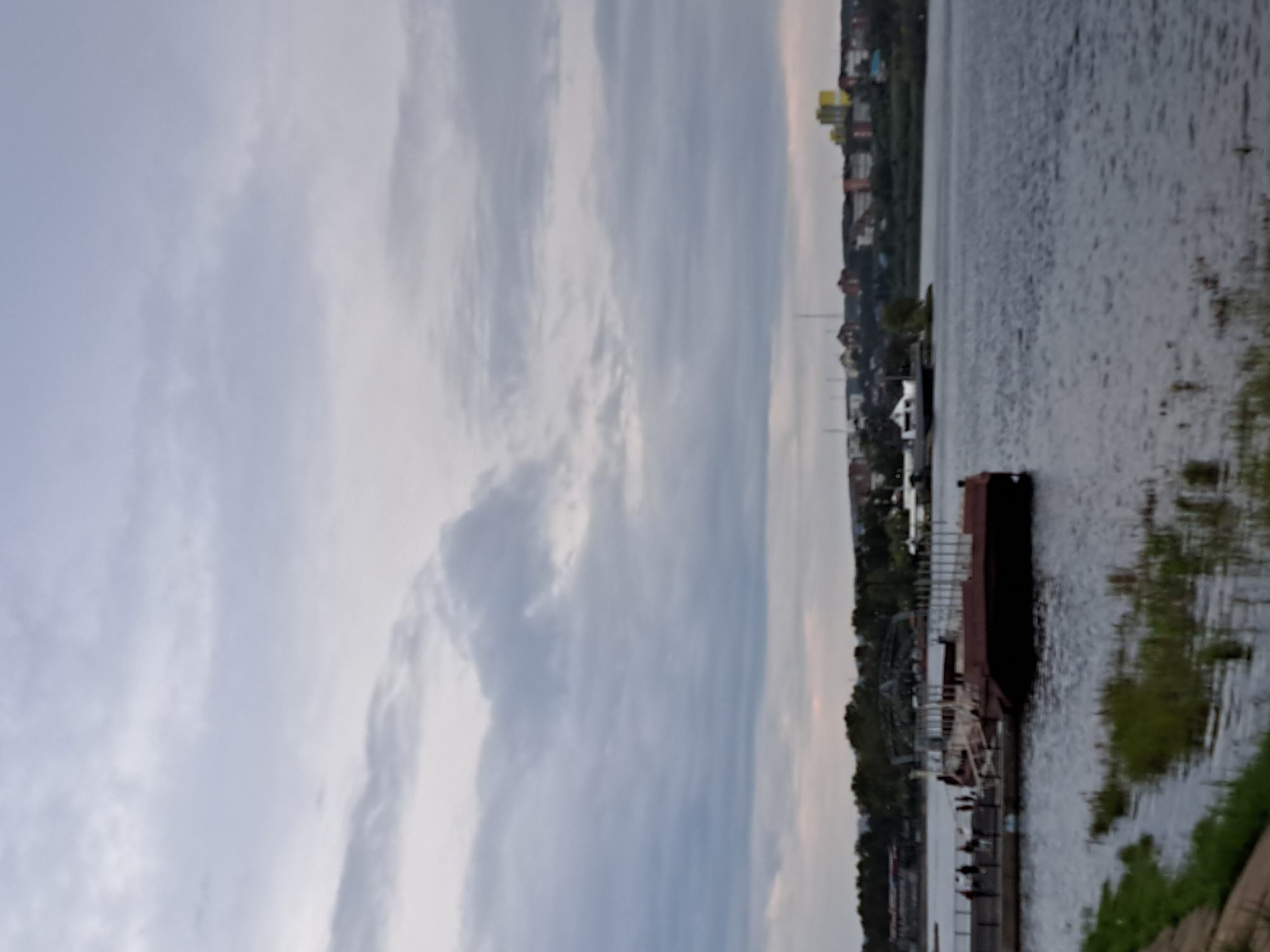 The image depicts a wide river with a barge and a pier in the foreground. Several people are visible standing on the pier, some appearing to be engaged in conversation. In the background, a cityscape with buildings and trees lines the far bank of the river. The sky is overcast with clouds. The overall scene suggests a view of the Angara River in Irkutsk, Russia, likely near a public access point or a small port.