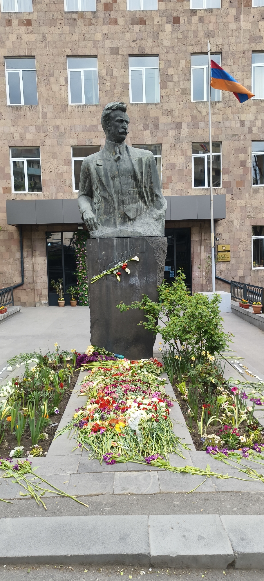 The photograph shows a bronze statue of Hovhannes Tumanyan in Yerevan, Armenia. The statue, placed in front of a brick building with multiple windows and a gray entrance, is adorned with fresh flowers. The Armenian flag is visible, situated next to the building entrance. Flowers are laid at the base of the statue, forming a colorful arrangement in the surrounding area.
