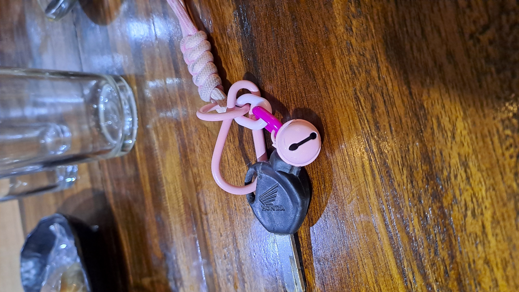 A close-up, eye-level shot shows a Honda key with a pink and white lanyard attached, resting on a dark wood grain surface. The key's black plastic head, bearing the Honda logo, is on the left. Attached to the key by a pink loop and a pink chain with two links is a small, light pink bell with a black clapper. The lanyard, made of braided pink and white cord, extends to the right, secured by a knot. In the background, out of focus, are two clear glass tumblers and a dark, shallow bowl containing food. The lighting suggests an indoor setting, possibly a restaurant or home. This scene does not provide any information regarding Pulokiton, Indonesia.