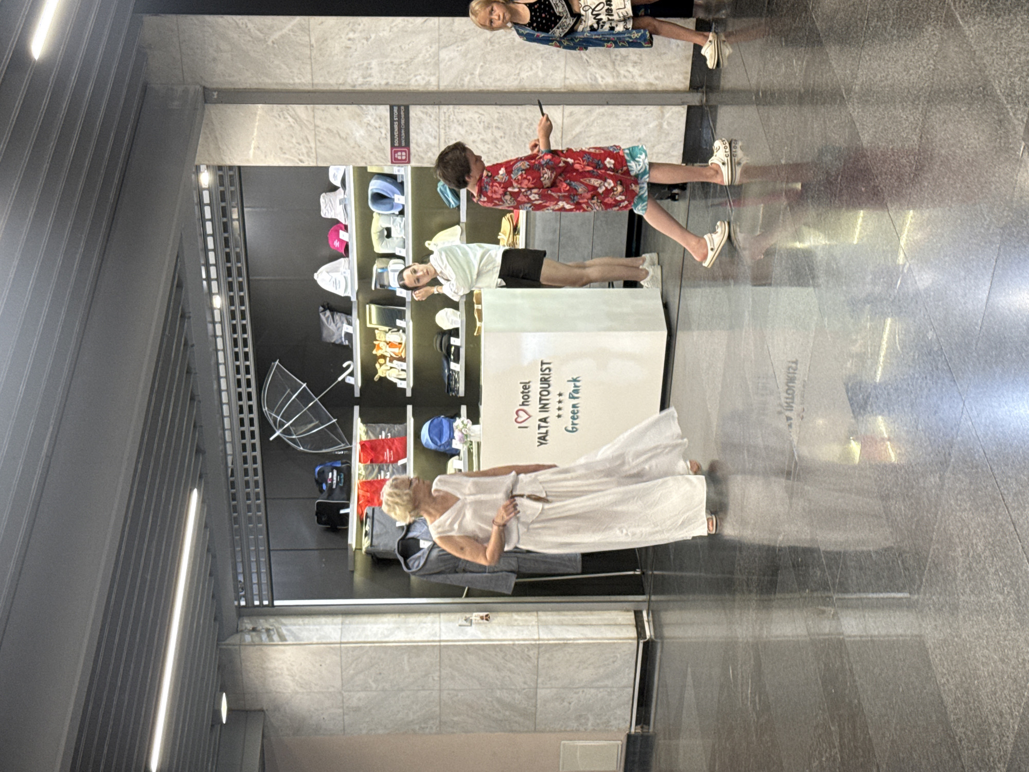 An indoor scene depicts a souvenir store within the Yalta Intourist Green Park hotel in Yalta, Ukraine. An older woman in a long white dress stands on the left, facing right. In the center, a young woman in a white top and black shorts stands behind a counter. To the right of the counter, a young boy wearing a red floral shirt and patterned shorts walks past, holding a mobile phone. Further to the right, a young girl with blonde hair in a black and blue patterned outfit stands. The store displays various items on shelves, including bags, hats, umbrellas, and what appear to be neck pillows. The floor is polished and reflective.