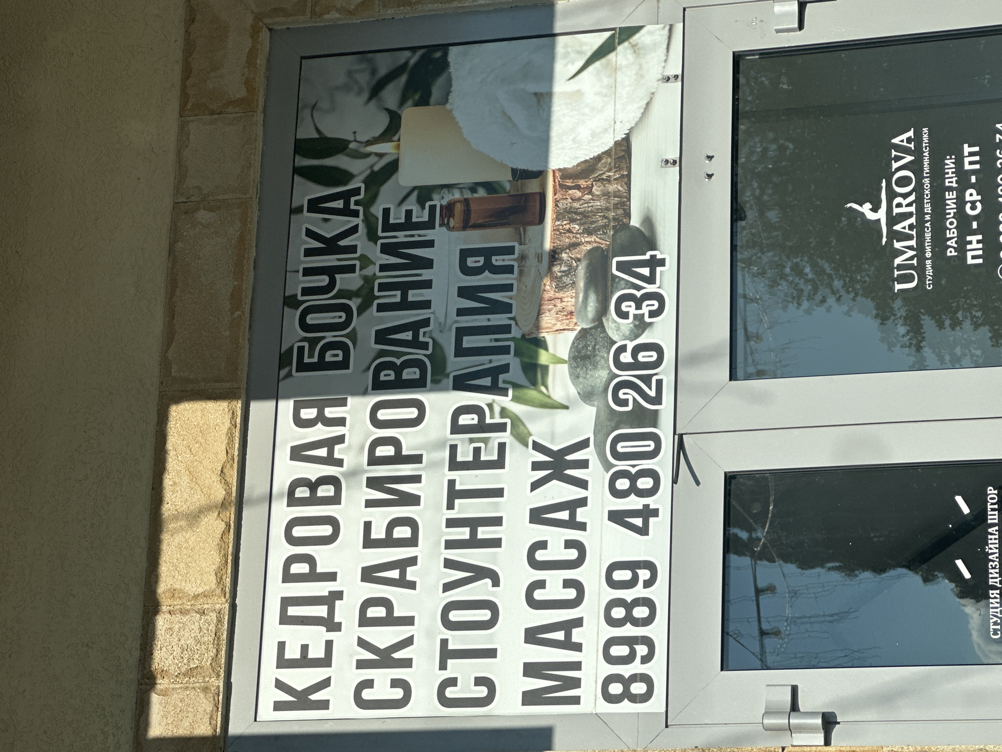 The image shows the facade of a building with a sign for a spa and fitness center. The sign, in large Cyrillic letters, advertises "CEDAR BARREL," "SCRUB," "STONE THERAPY," and "MASSAGE." Below this text is a phone number: "8989 480 26 34." The sign also features an image of spa elements, including candles, stones, and a white towel.
Below the main sign, partially visible through a glass door, is a logo and text for "UMAROVA," a fitness and children's gymnastics studio. This text indicates operating days: "MON - WED - FRI." The building appears to be in Miatli, Russia, as indicated by the location context. The overall scene is a commercial storefront with signage for health and wellness services.