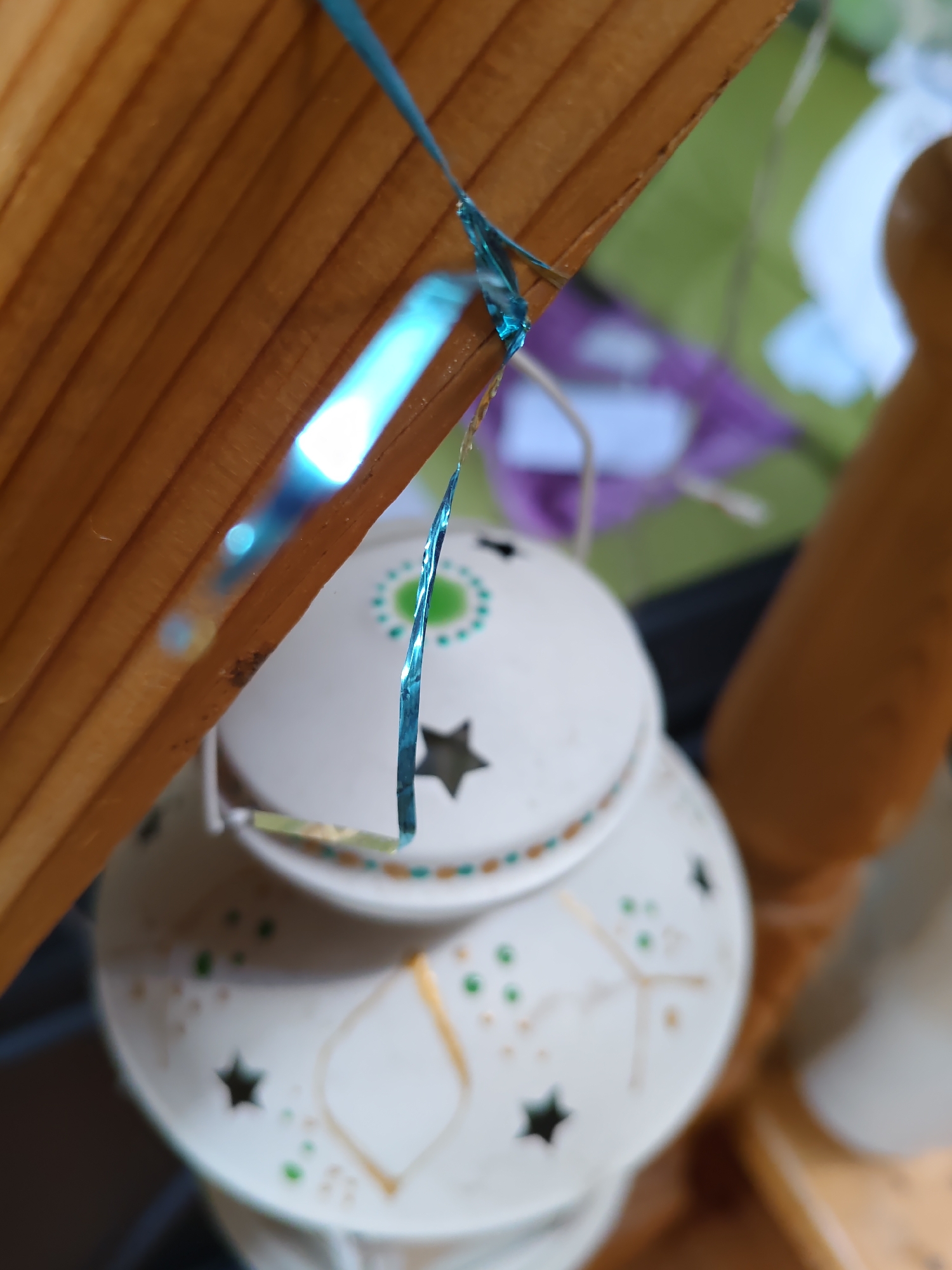 The image shows a close-up of decorative elements, possibly part of a festive or themed display. A wooden structure, with horizontal grain, dominates the upper left portion of the frame. A thin, iridescent blue ribbon is draped across this structure and extends downwards. Attached to the ribbon, and partially visible, is a white, lantern-like object with decorative patterns. These patterns include small green dots, gold-colored lines forming geometric shapes, and cut-out star shapes. The lantern appears to be made of metal or ceramic and has a lid with a circular pattern featuring green and white accents. The background is softly blurred, showing hints of green and purple, suggesting an outdoor or indoor setting with other decorative items. The lighting in the scene creates reflections and highlights on the ribbon and the wooden surface. There are no people visible in the image. The image does not contain any information that would indicate its location in Rostov-on-Don, Russia.