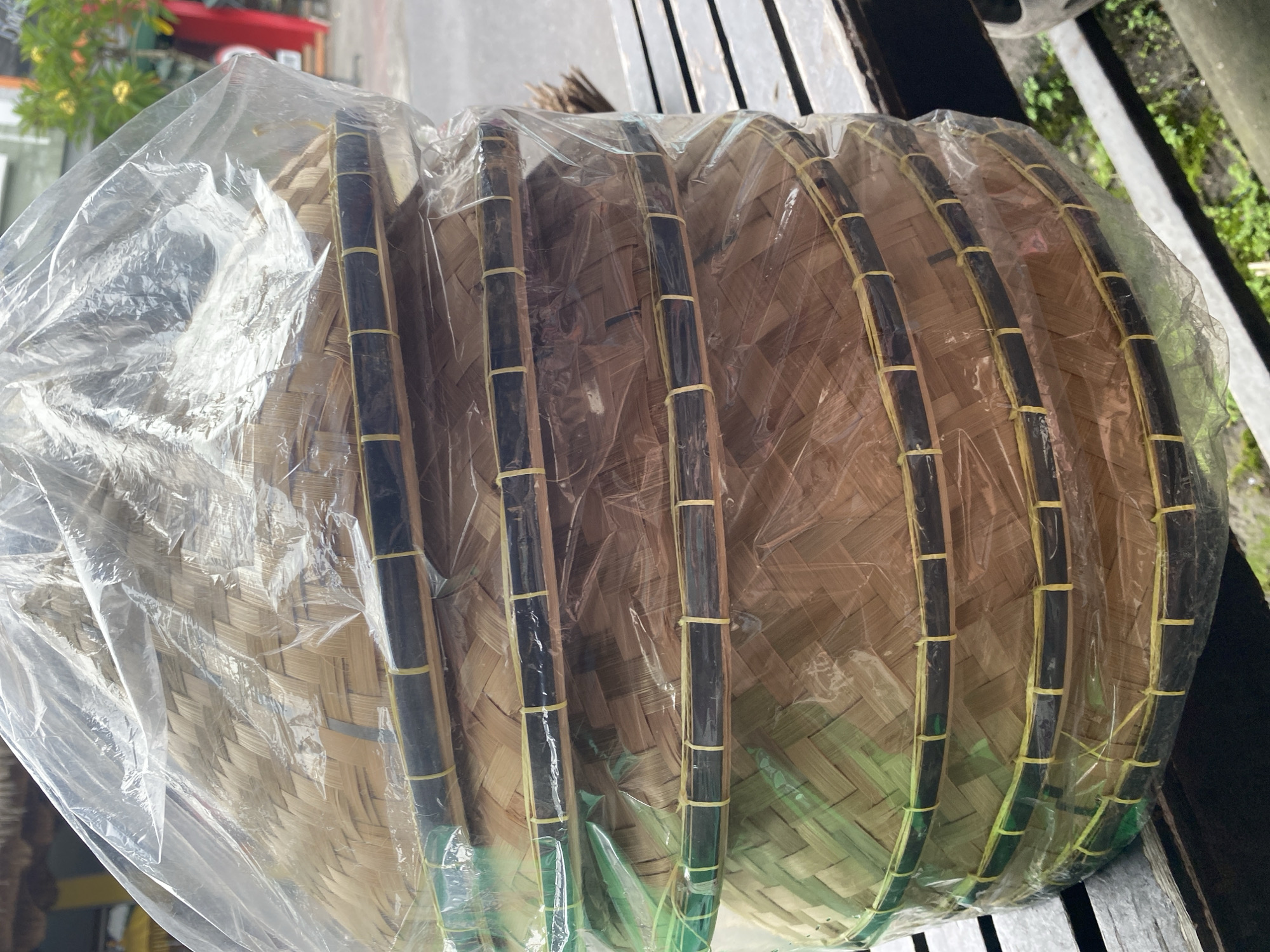 A stack of six round woven trays with dark rims, secured by thin, light-colored bands, is packaged in clear plastic. The trays rest on a weathered wooden surface with slats. In the background, there are glimpses of greenery, parts of buildings, and possibly a vehicle, suggesting an outdoor location in Kuta Selatan, Indonesia.