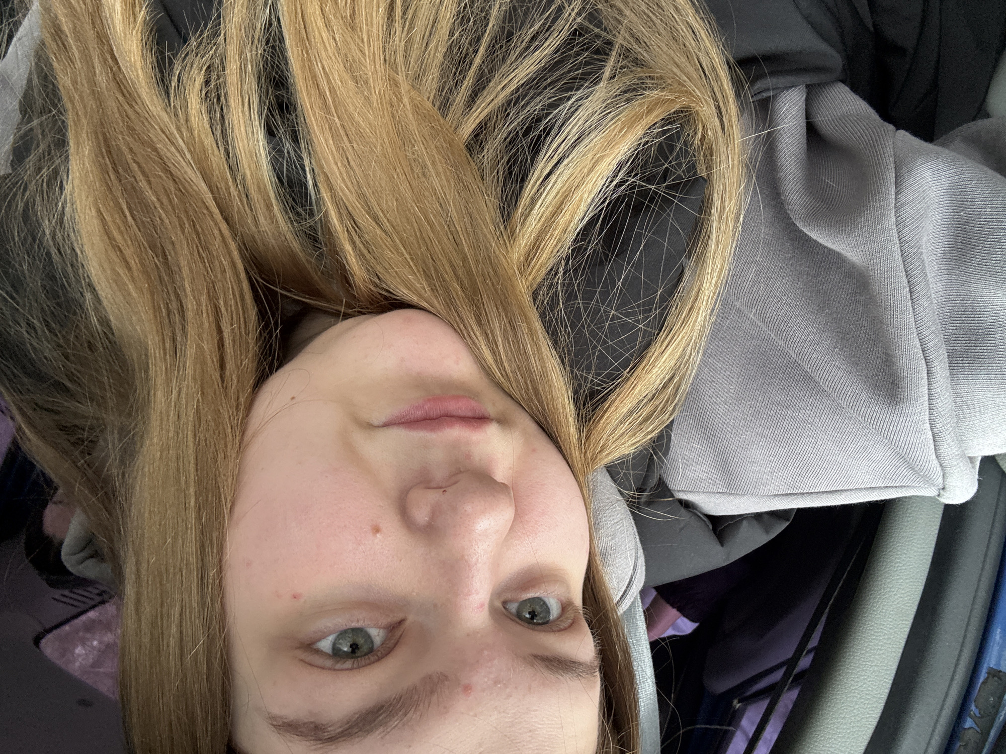 A medium shot depicts a Caucasian female with light brown hair, tilted to the side. Her eyes are grey, and she has light skin with some blemishes. She is wearing a grey hoodie. The background shows a car interior with parts of the dashboard, door panel, and seat visible. The image does not reveal any information about the location being Chaikovsky, Russia.