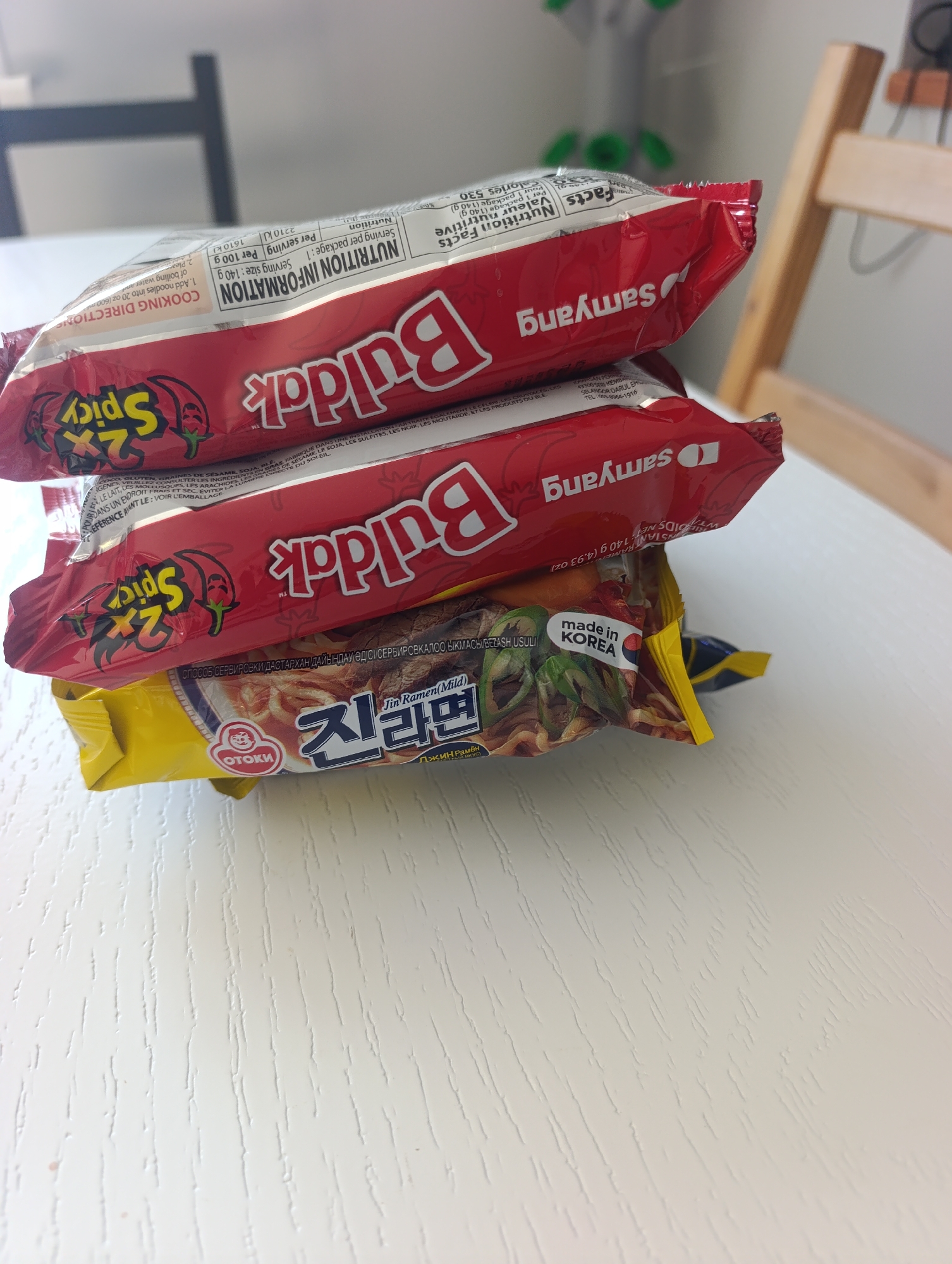 A stack of instant ramen noodle packages sits on a white wooden table. The top two packages are red with the brand name "Buldak" in large white lettering and "Samyang" in smaller white lettering. Both packages have a "2X Spicy" graphic with chili peppers. The third package from the top is also red with "Samyang" branding and "Buldak" labeling. The bottom package is yellow and features Korean text that appears to say "Jin Ramen" and "OTOKI". A small circle with a face is visible on the yellow package, and the text "made in KOREA" is also present. The background is blurred but suggests an indoor setting with furniture. The image is taken from a slightly elevated angle, looking down at the stack of ramen. There are no people or animals visible in the image. The location is not identifiable as Ivanovca, Russia.
