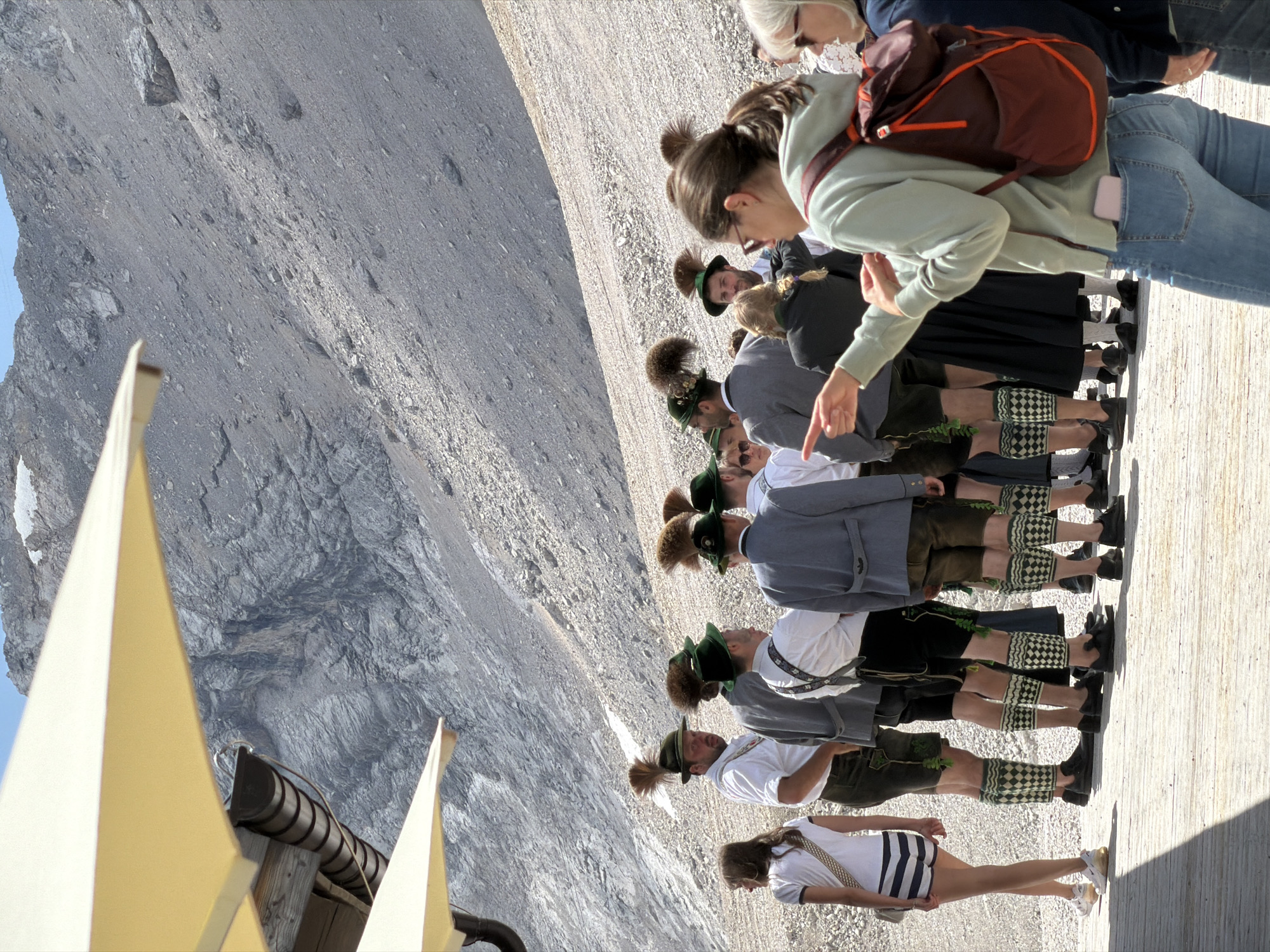 This image was taken at the Zugspitze, Germany's highest peak, near Garmisch-Partenkirchen. A group of people are gathered on a wooden platform. Several men are dressed in traditional Bavarian attire, including lederhosen, suspenders, white shirts, and feathered hats. They appear to be posing or waiting for a tour. A young woman in a t-shirt and striped shorts stands to the left. In the foreground, a woman in a light green hoodie and jeans points to the left, wearing a maroon backpack. The background shows a steep, rocky mountainside under a bright, clear sky, with the suggestion of snow or ice patches higher up. Yellow awnings provide shade in the upper left corner.
