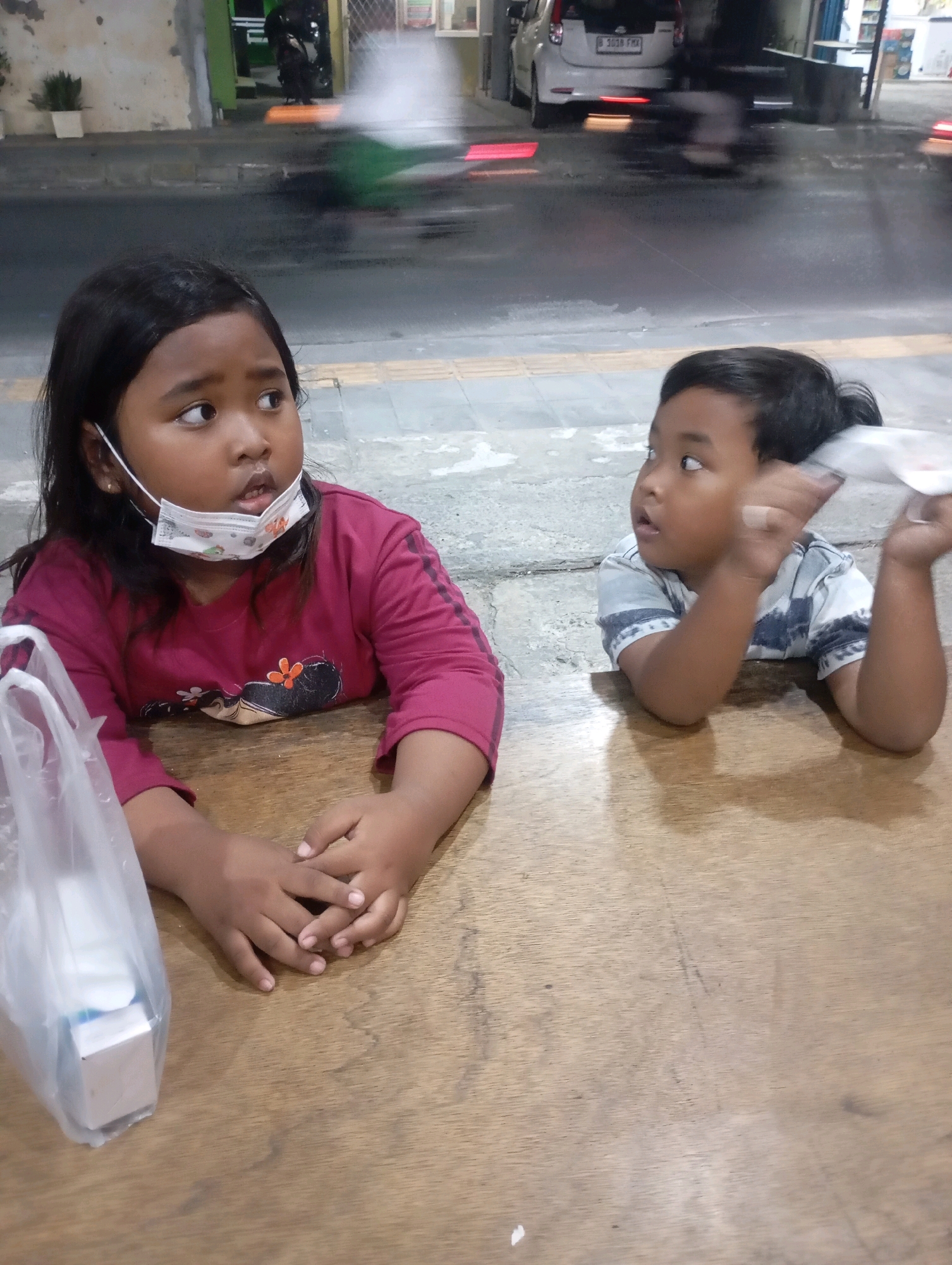 The image shows two young children, a girl and a boy, sitting at a wooden table. The girl, on the left, has long dark hair and is wearing a maroon long-sleeved shirt. She has a white face mask with a cartoon pattern on it, pulled down below her nose. Her hands are clasped in front of her. To her right, the boy is wearing a striped t-shirt and is looking to his right, holding a small white object in his right hand. A clear plastic bag containing a white box is on the table to the left of the girl. In the background, a street scene is visible with blurry vehicles and buildings, suggesting an outdoor or semi-outdoor setting. The environment appears to be a public area in Babelan, Indonesia, indicated by a license plate on a car in the background which reads "B 3039 FRX", a typical Indonesian plate format. The lighting suggests it is either dusk or nighttime.