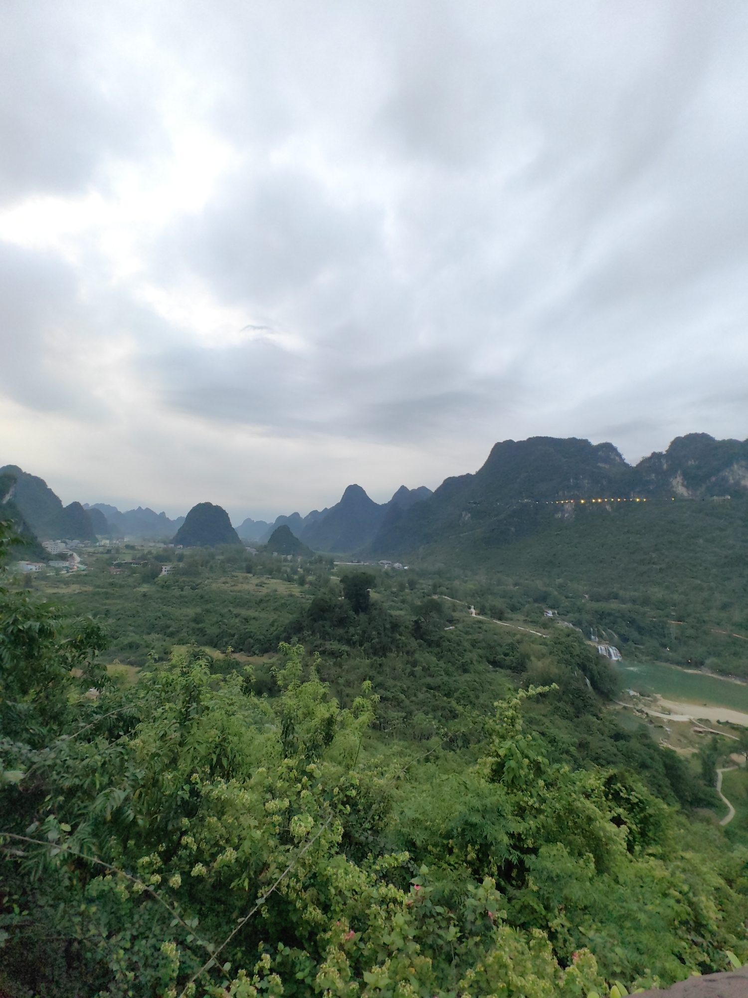 The image presents a wide-angle view of a landscape in Xã Đàm Thủy, Vietnam, under a cloudy sky. The scene is dominated by a series of green, karst mountains in the middle ground, diminishing in size and clarity with distance. A village nestled in the valley is visible in the center, with buildings scattered amidst greenery. The foreground consists of dense foliage and trees. A body of water is seen to the right, with a winding path adjacent to it. On the ridge of one of the mountains, lights are distributed along what appears to be a road or elevated structure.