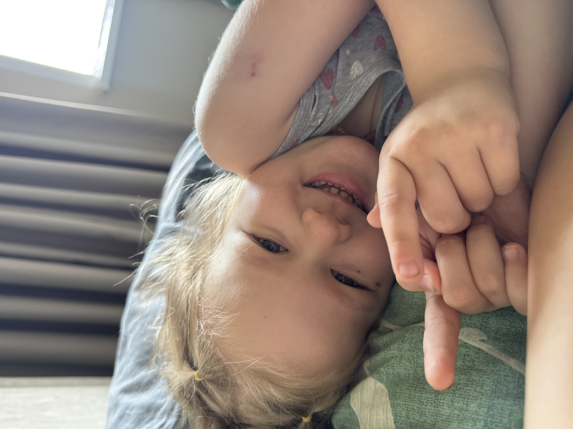 A close-up, rotated image depicts a young Caucasian child with blonde hair smiling and looking at the camera. The child is lying down, and their head is positioned near the bottom of the frame, with the image rotated such that the top of the head is oriented downwards. The child has fair skin and light blue eyes. They are wearing a gray shirt with red hearts on it. The child's right arm is raised, and their hand is clenched, with their index finger extended down. A portion of a parent/guardian's arm is visible on the right side of the image. The child and adult are both laying on a dark gray and a green and white patterned bed. In the background, a window with horizontal blinds is seen. The location is Ferma, Russia, based on the query.