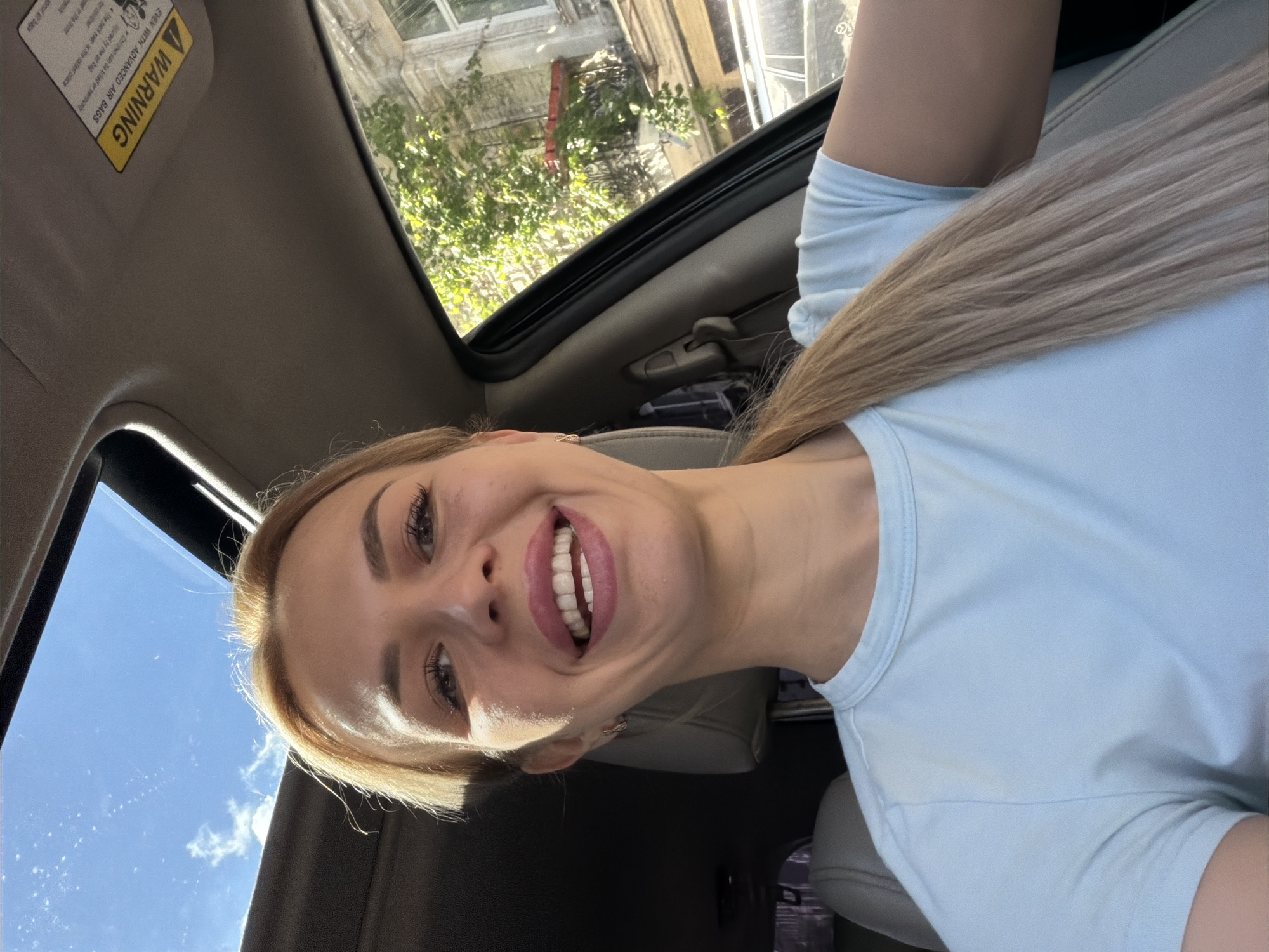 In a bright, sunlit selfie taken from inside a car, a woman with blonde hair pulled back in a ponytail smiles broadly. She is wearing a light blue t-shirt. Through the open sunroof, a clear blue sky with scattered clouds is visible. The interior of the car shows a warning sticker on the sun visor and the back of a car seat. Through the car window, a tree with green leaves and part of a building with ornate architectural details can be seen, suggesting an urban setting. The background and architecture are consistent with the historical districts of Kyiv, Ukraine.